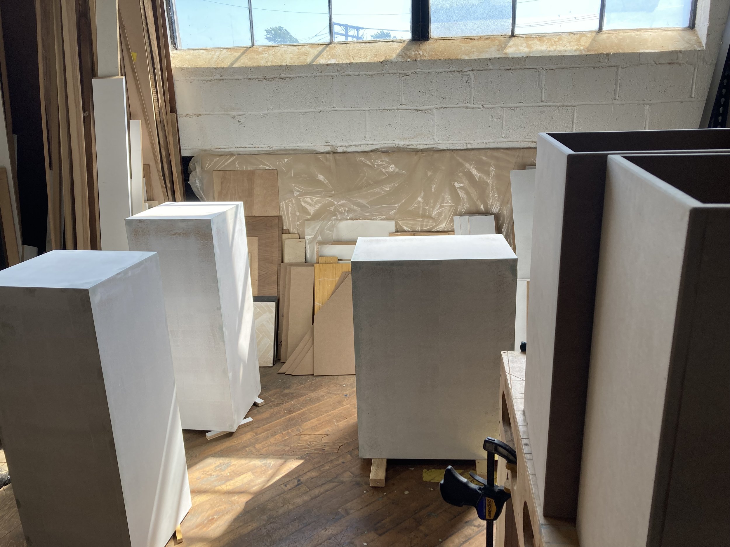 Woodworking workshop with large white wooden blocks, various wooden panels, clamps, and natural light coming through a window.