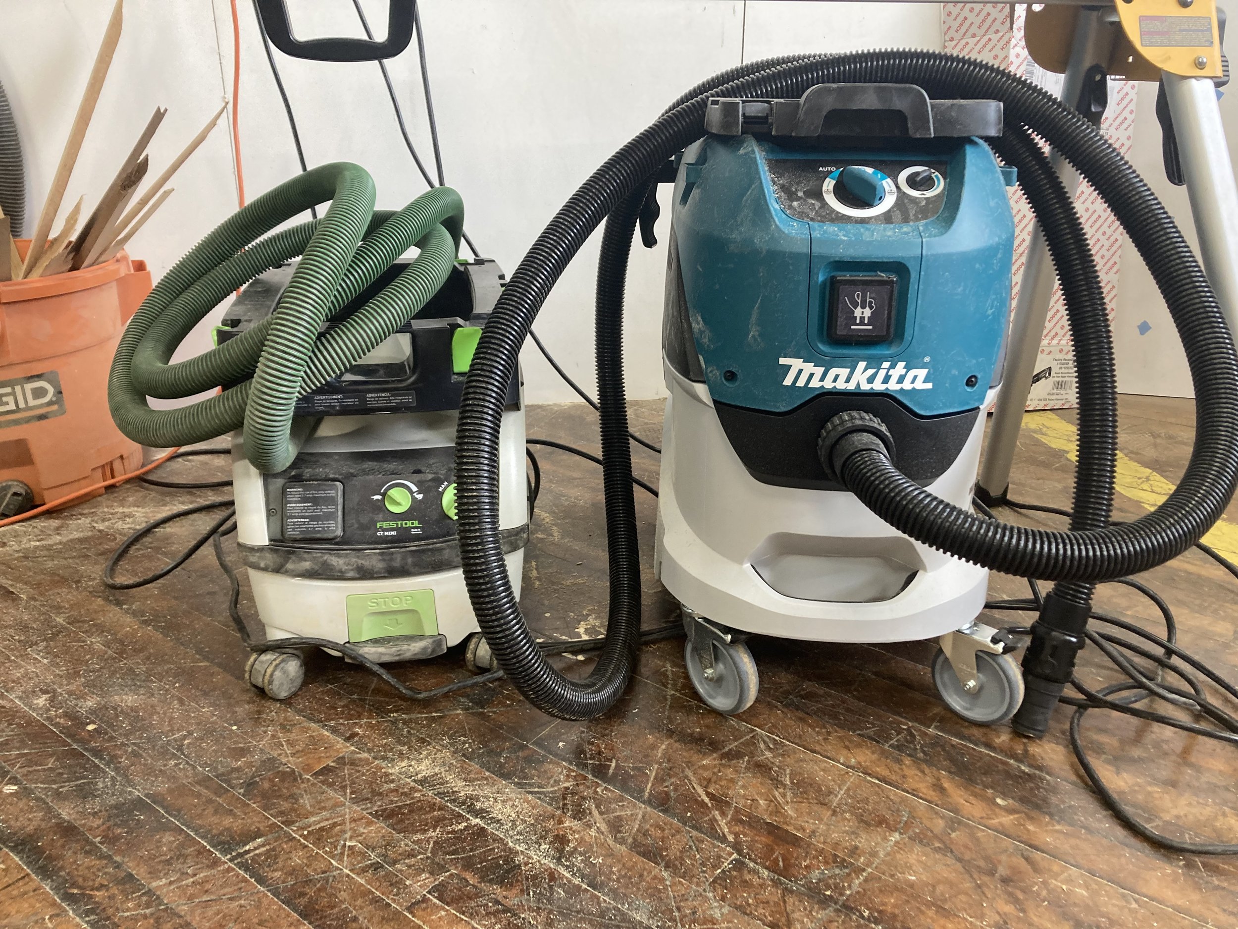 Two shop vacuums, one Festool with a green hose and one Makita with a black hose, on a workshop floor.