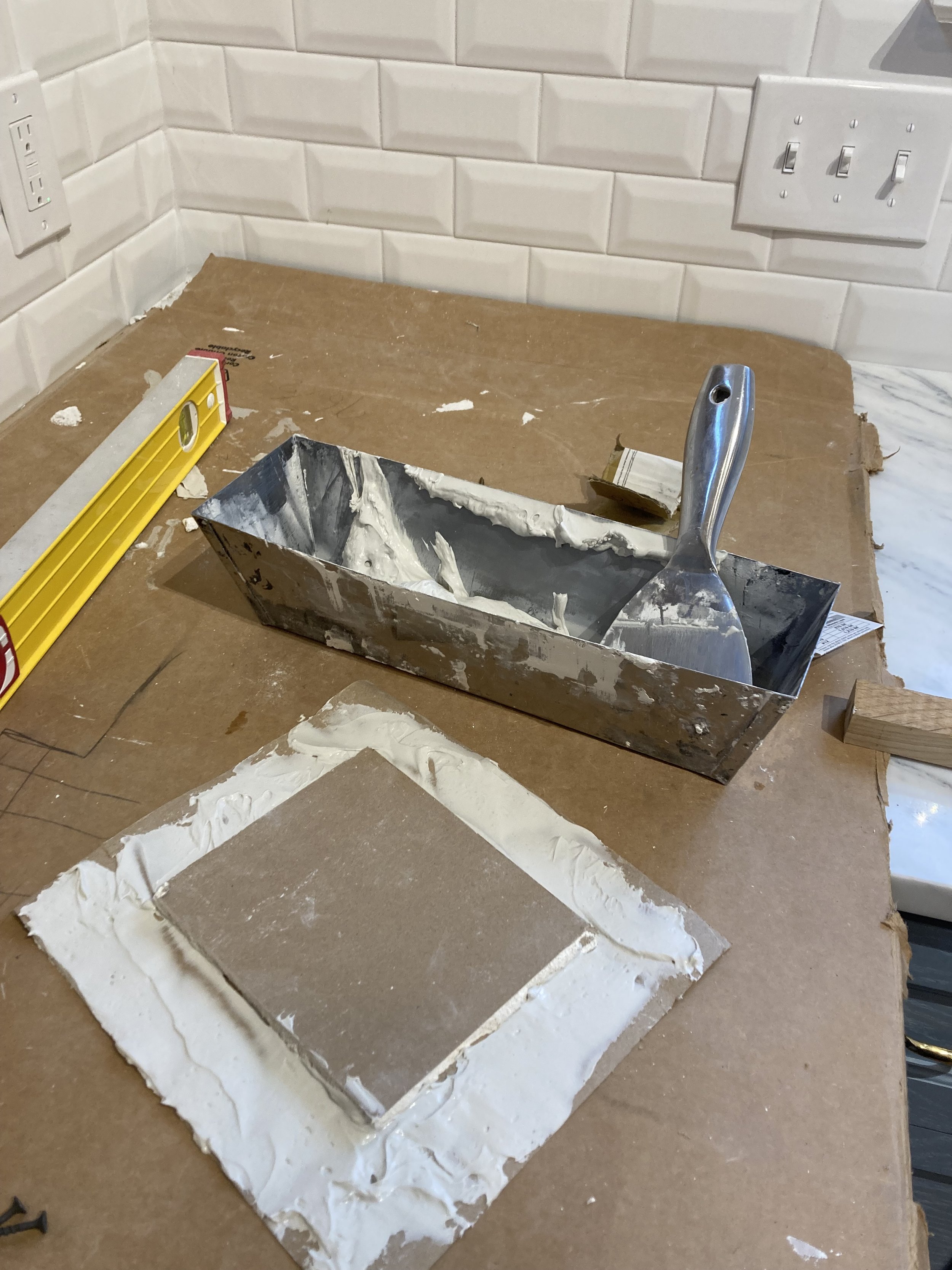 Construction tools on a table, including a putty knife, metal container with joint compound, yellow level, and a tile covered in adhesive, against a tiled backsplash.