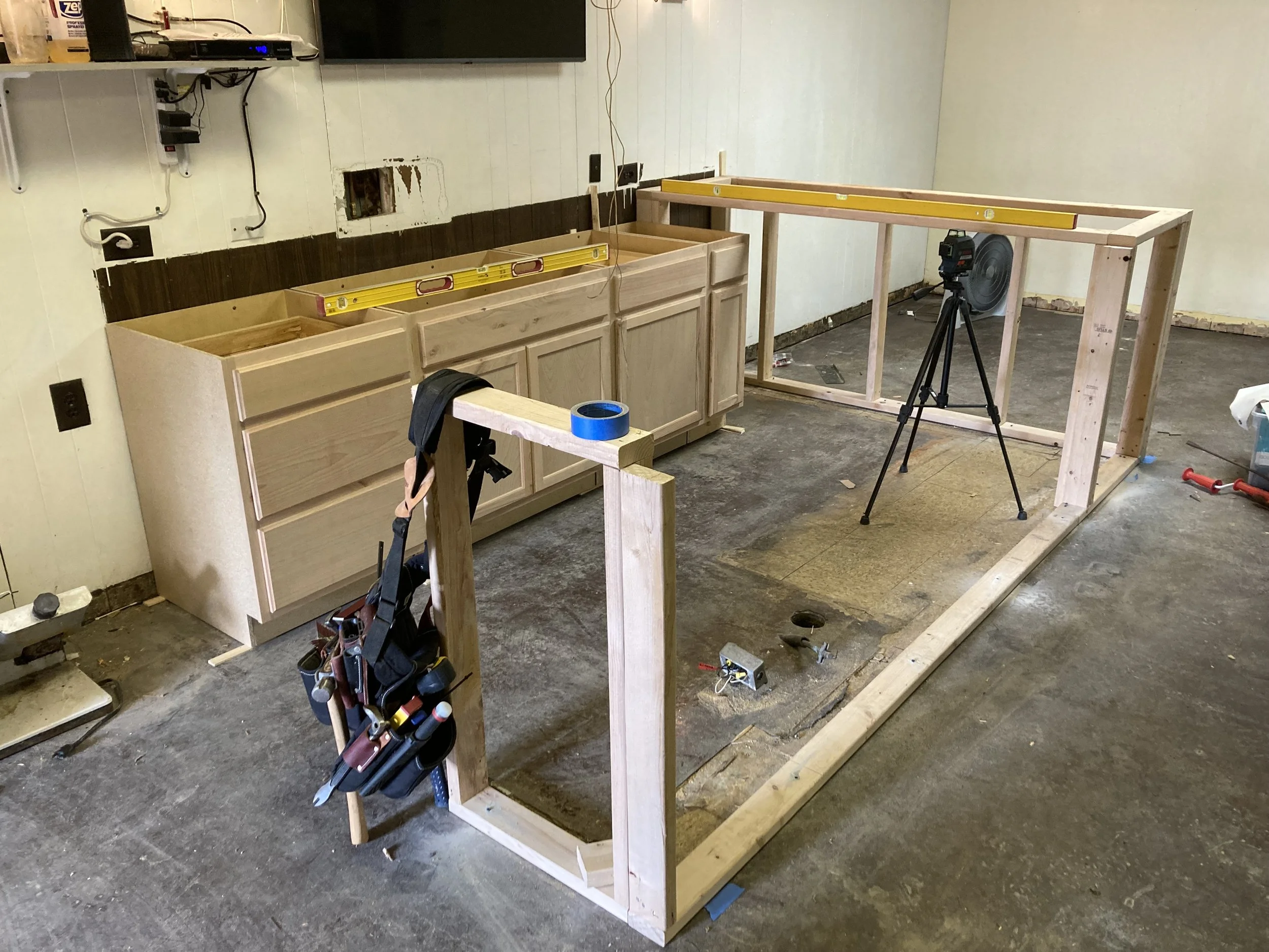 Kitchen renovation scene with partially installed cabinets, a workbench frame, construction tools, tripod with laser level, and unfinished wall.