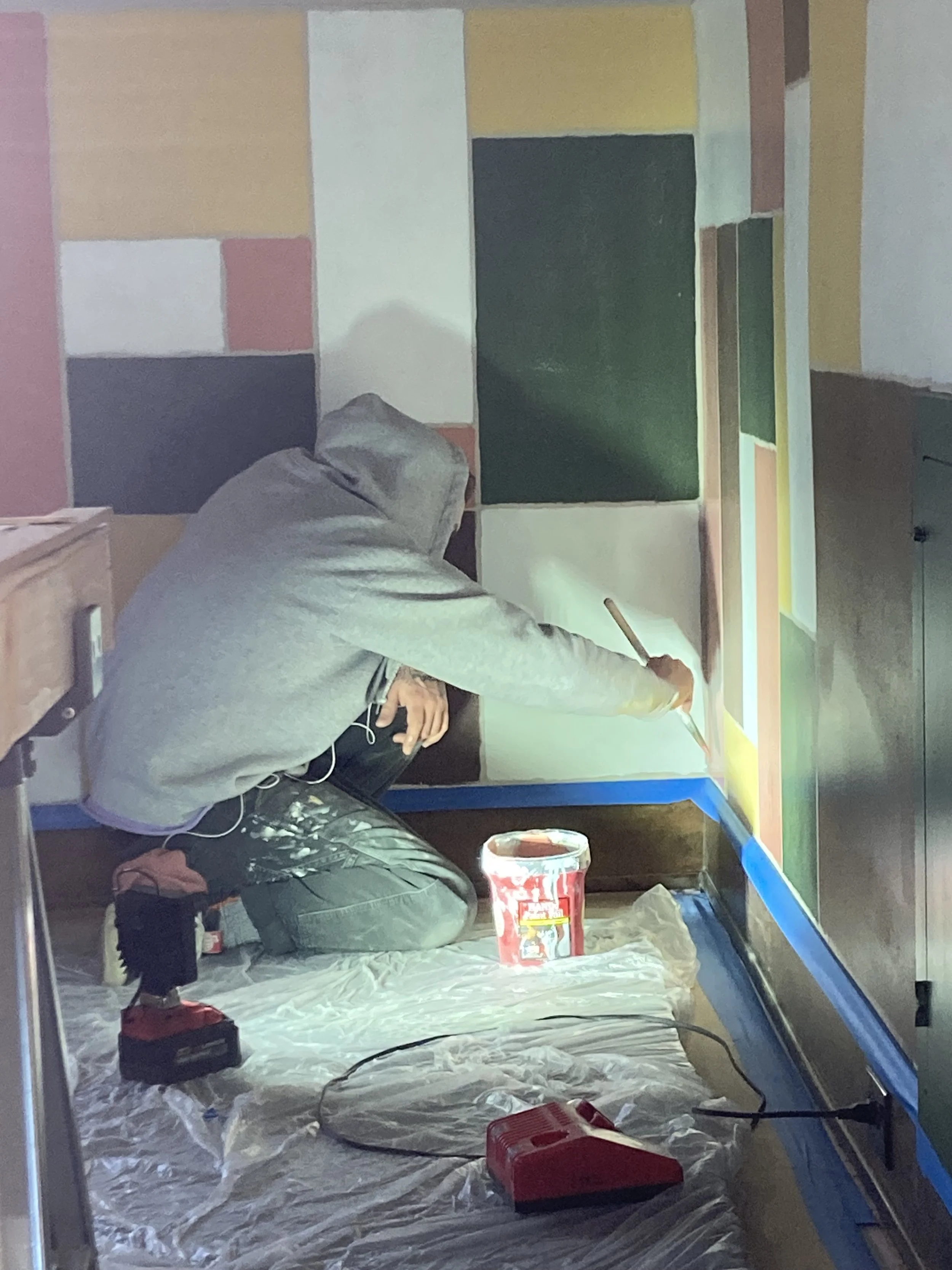 Person painting a wall with geometric shapes, wearing grey hoodie and black pants, surrounded by painting supplies.