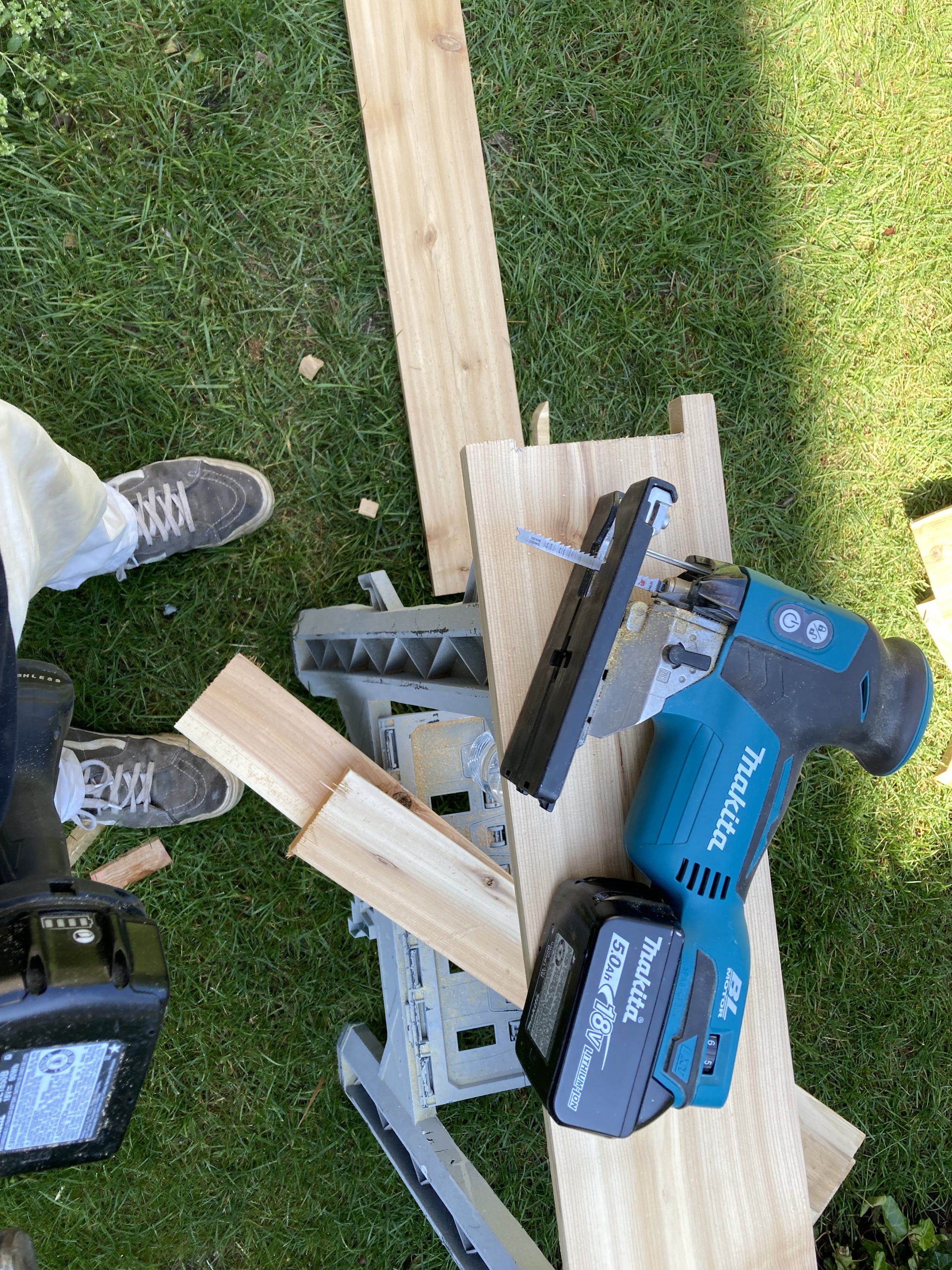 A cordless jigsaw on a piece of wooden plank, surrounded by other wooden pieces and a saw horse on grass.