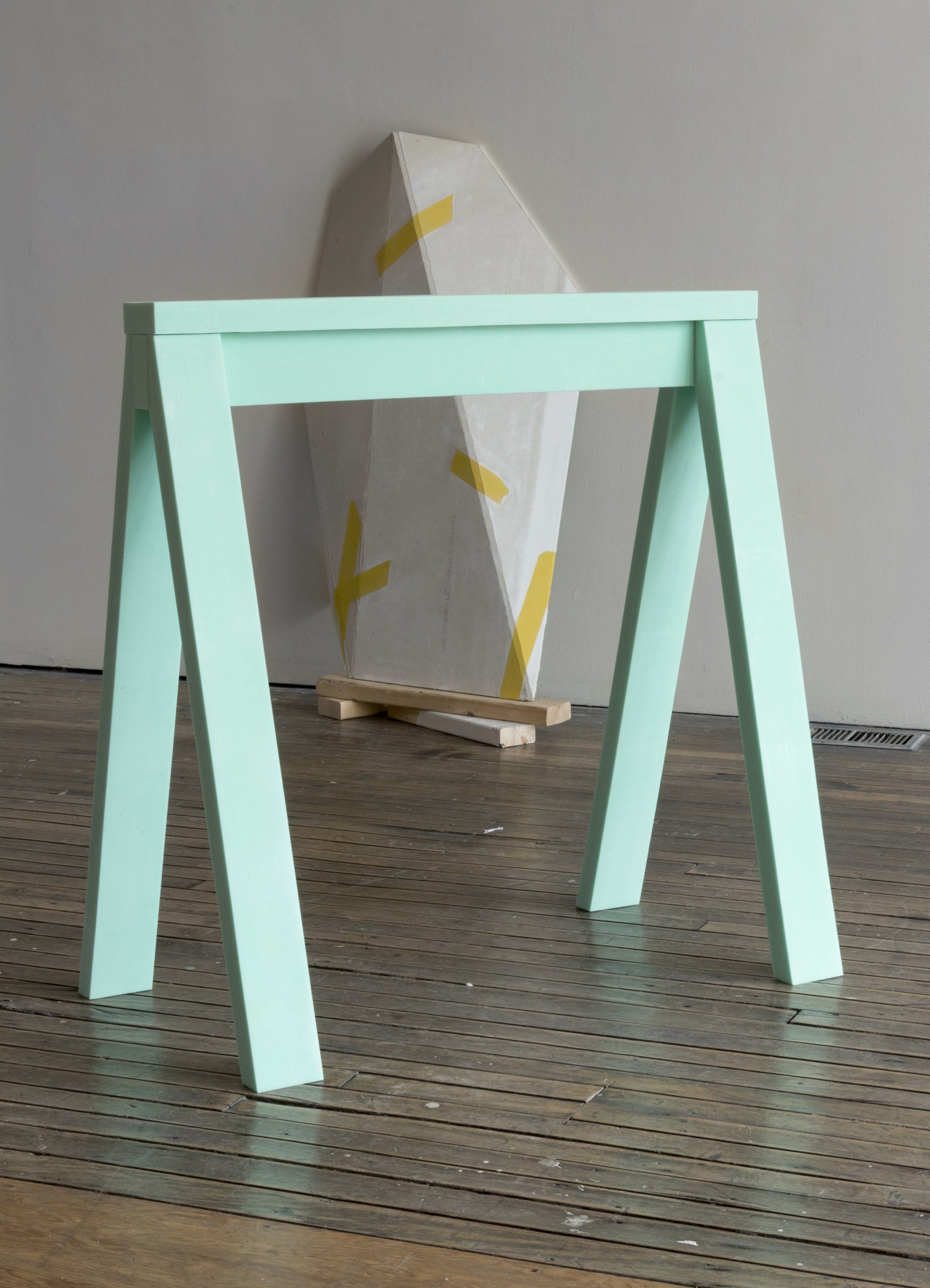 Mint green wooden sawhorse on a wooden floor with a geometric art piece in the background.