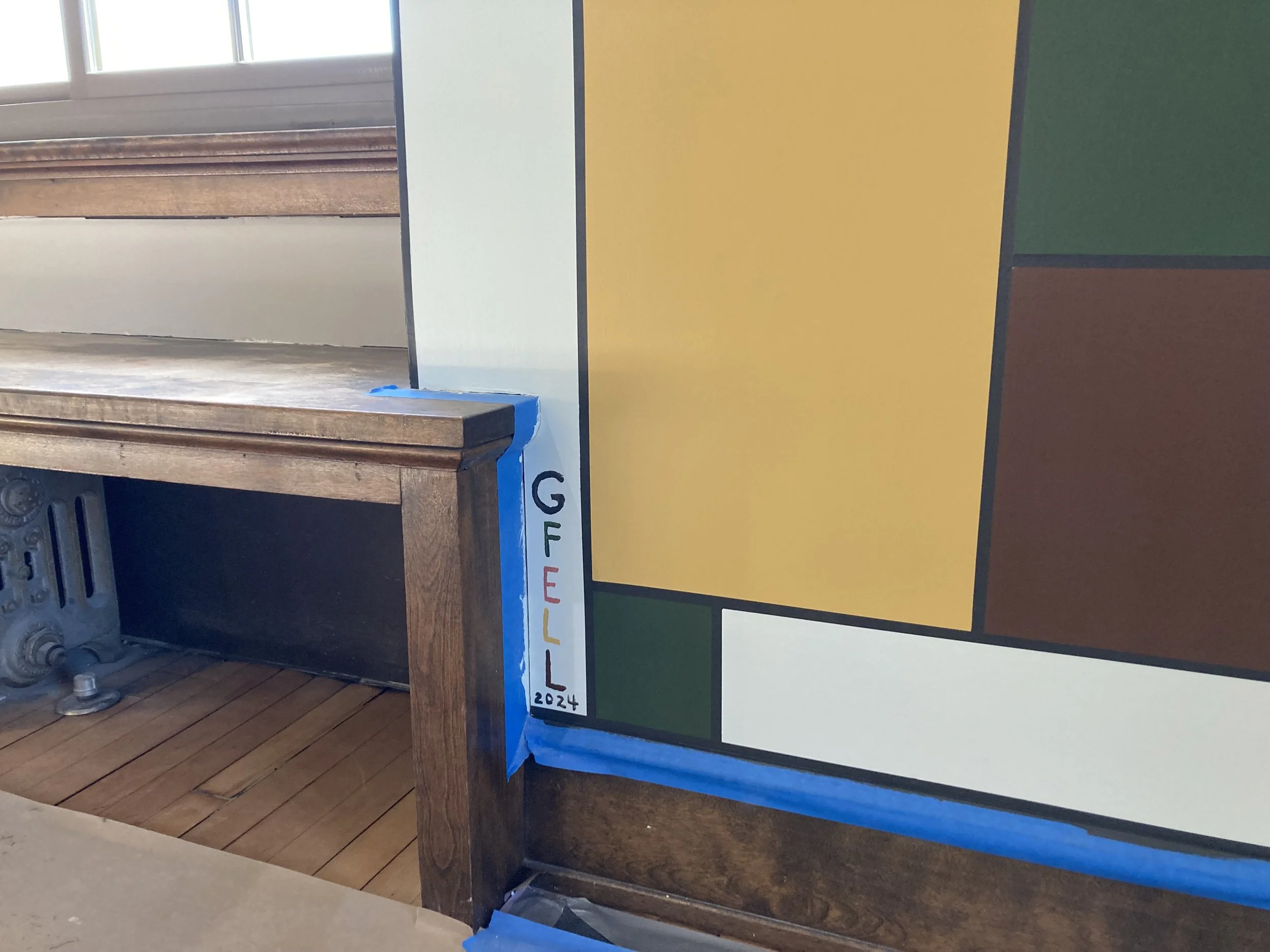 Wooden bench next to abstract geometric painting with yellow, green, and brown blocks, blue painter's tape, radiator underneath, and "GFELL 2024" written on the edge.