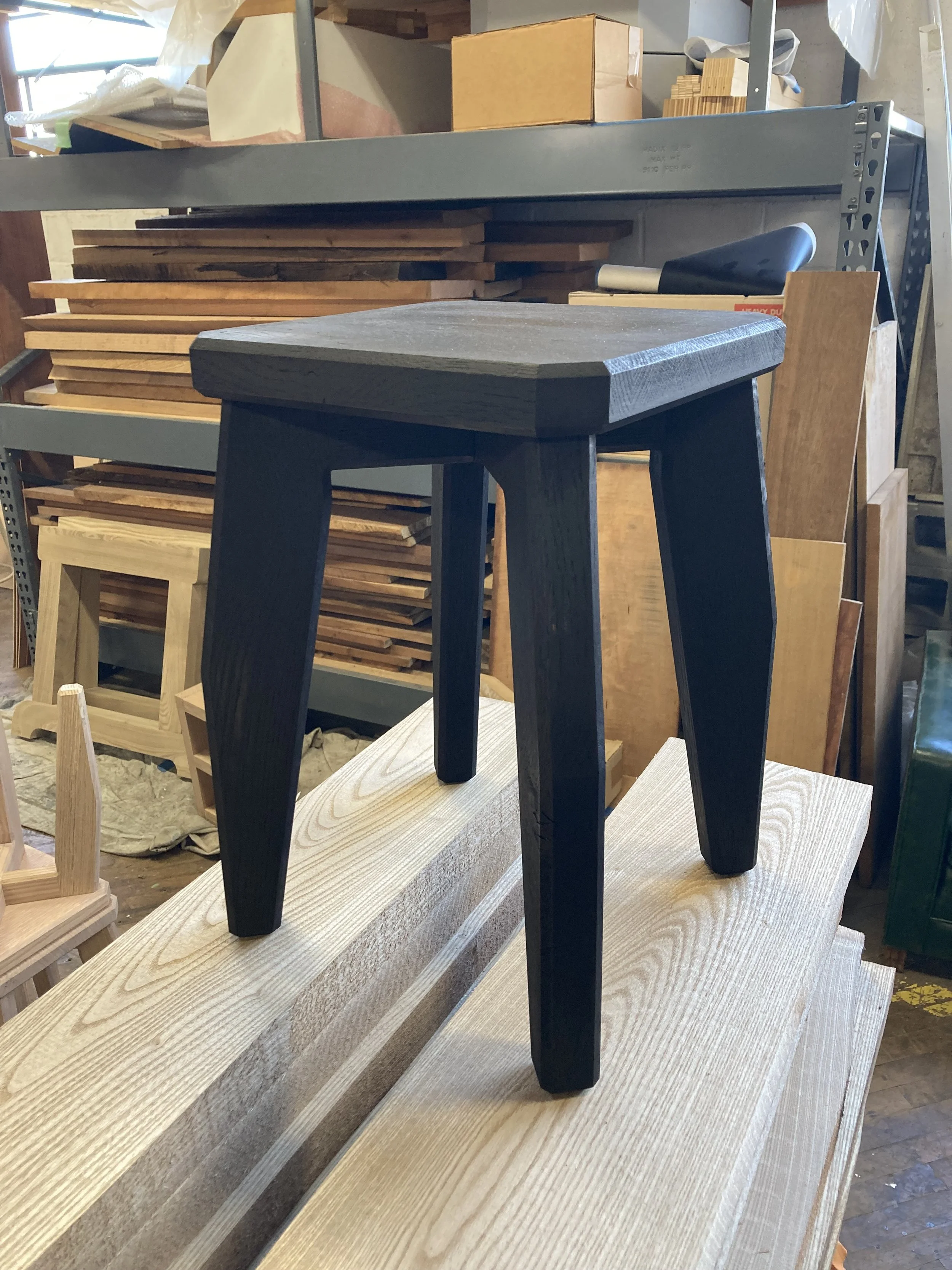 A black wooden stool placed on stacked wooden planks in a workshop with shelving in the background.