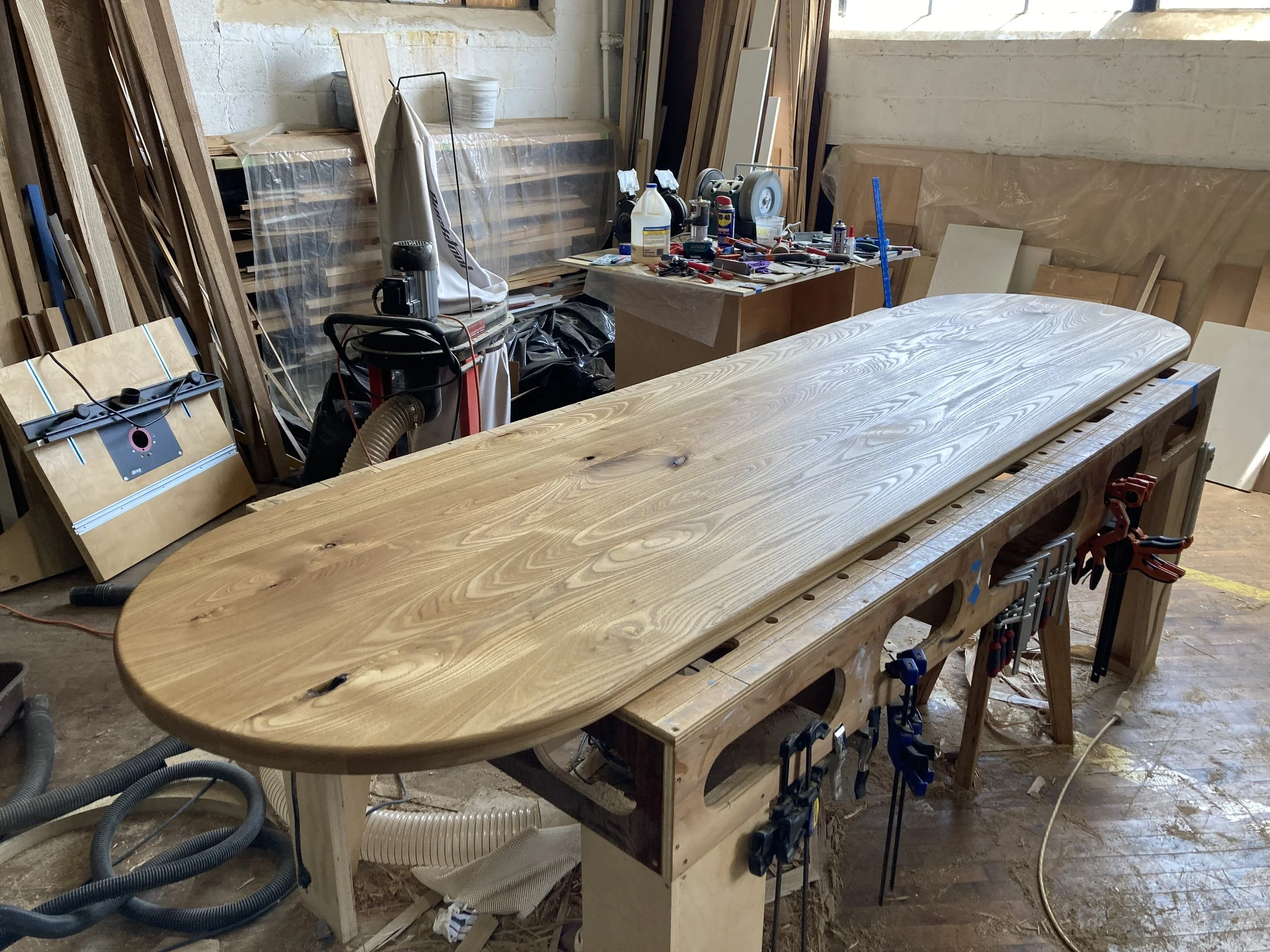 Wooden surfboard on a workbench in a woodworking shop with tools and materials in the background.