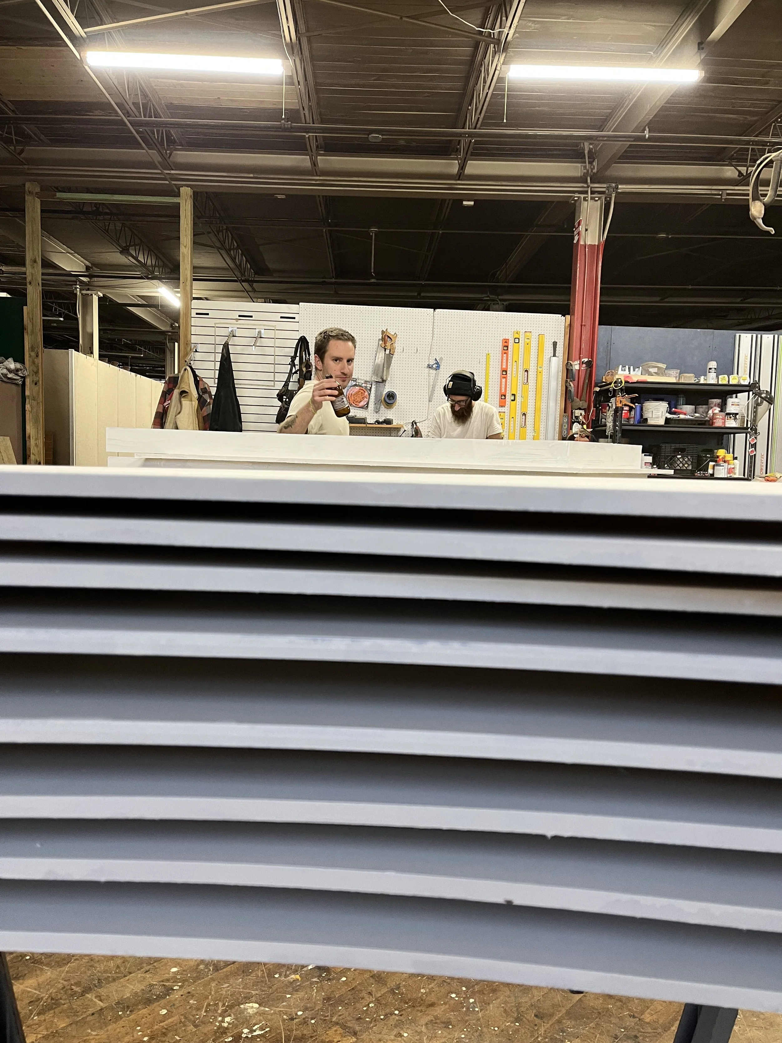 Two people working in a workshop with tools and supplies visible, focusing on stacked sheets of material in the foreground.