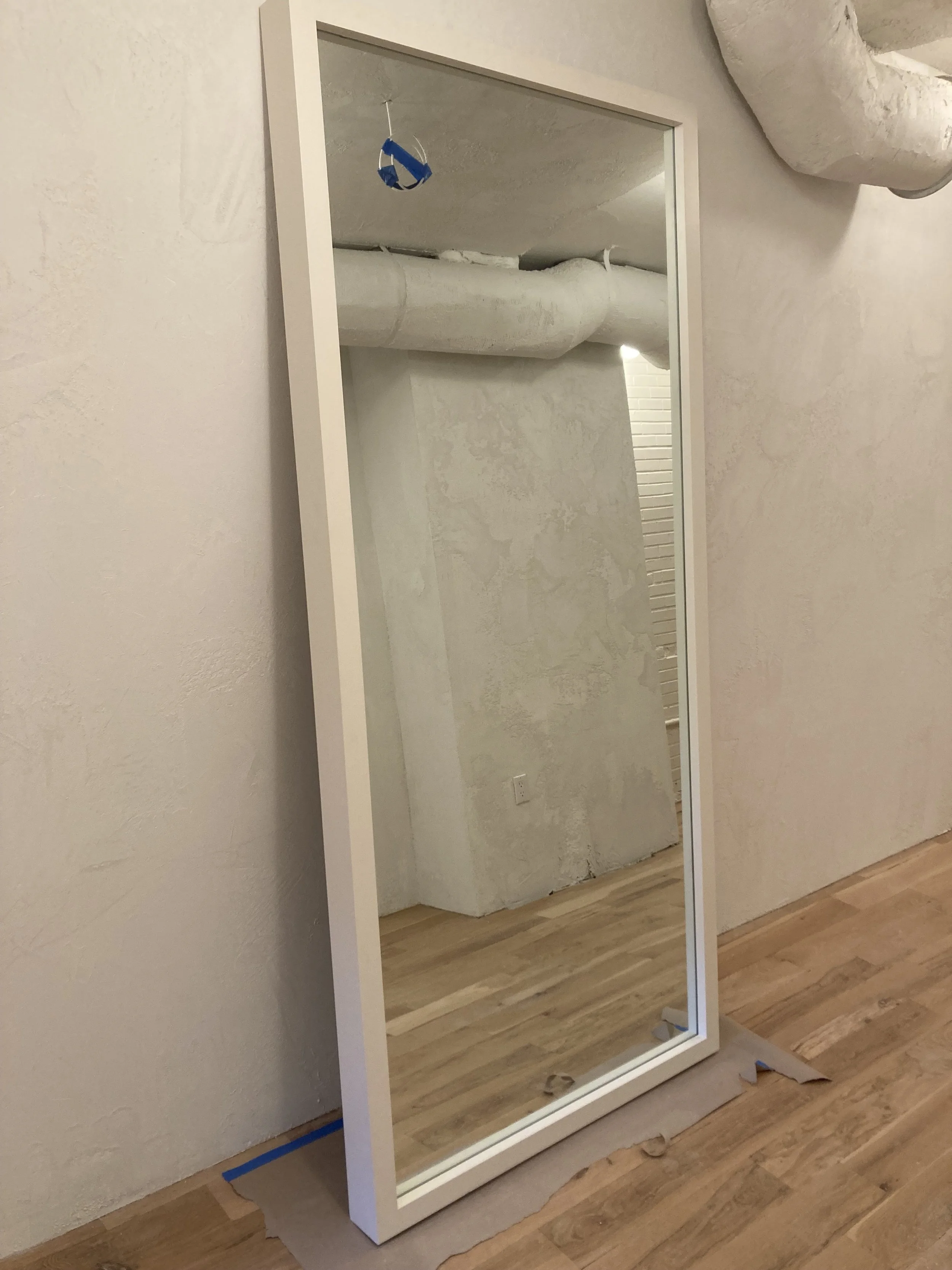 Full-length mirror with a white frame leaning against a textured wall in a room with wood flooring and exposed ductwork.