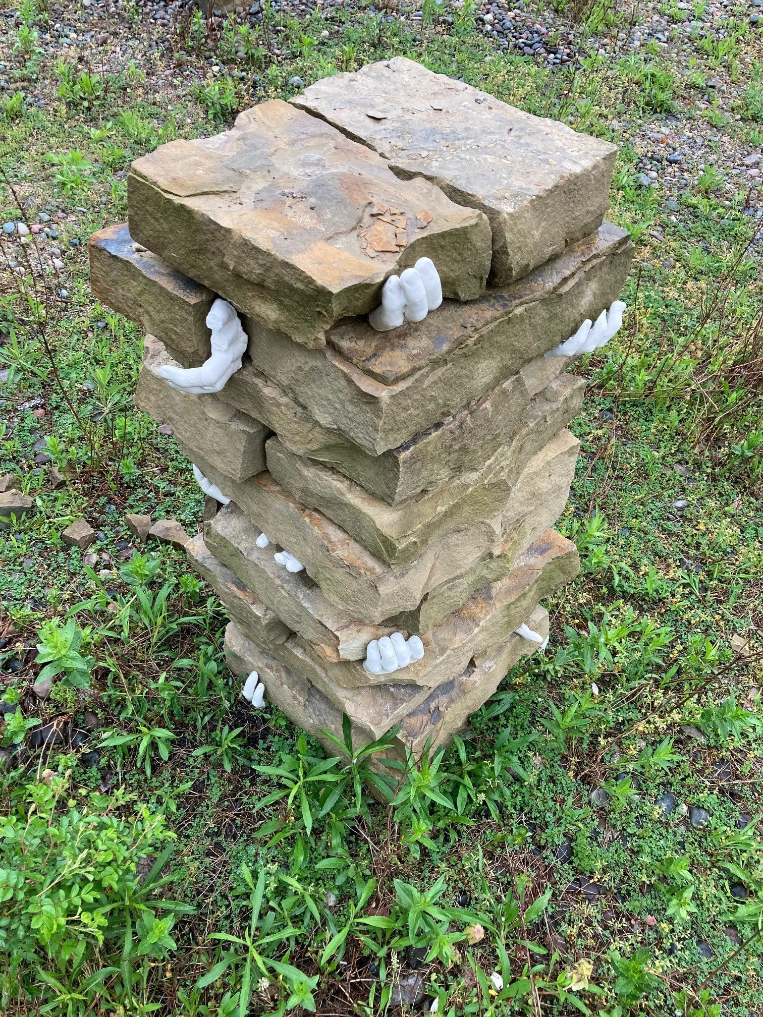 Stack of flat stones with white hand sculptures between layers, set on grassy ground.