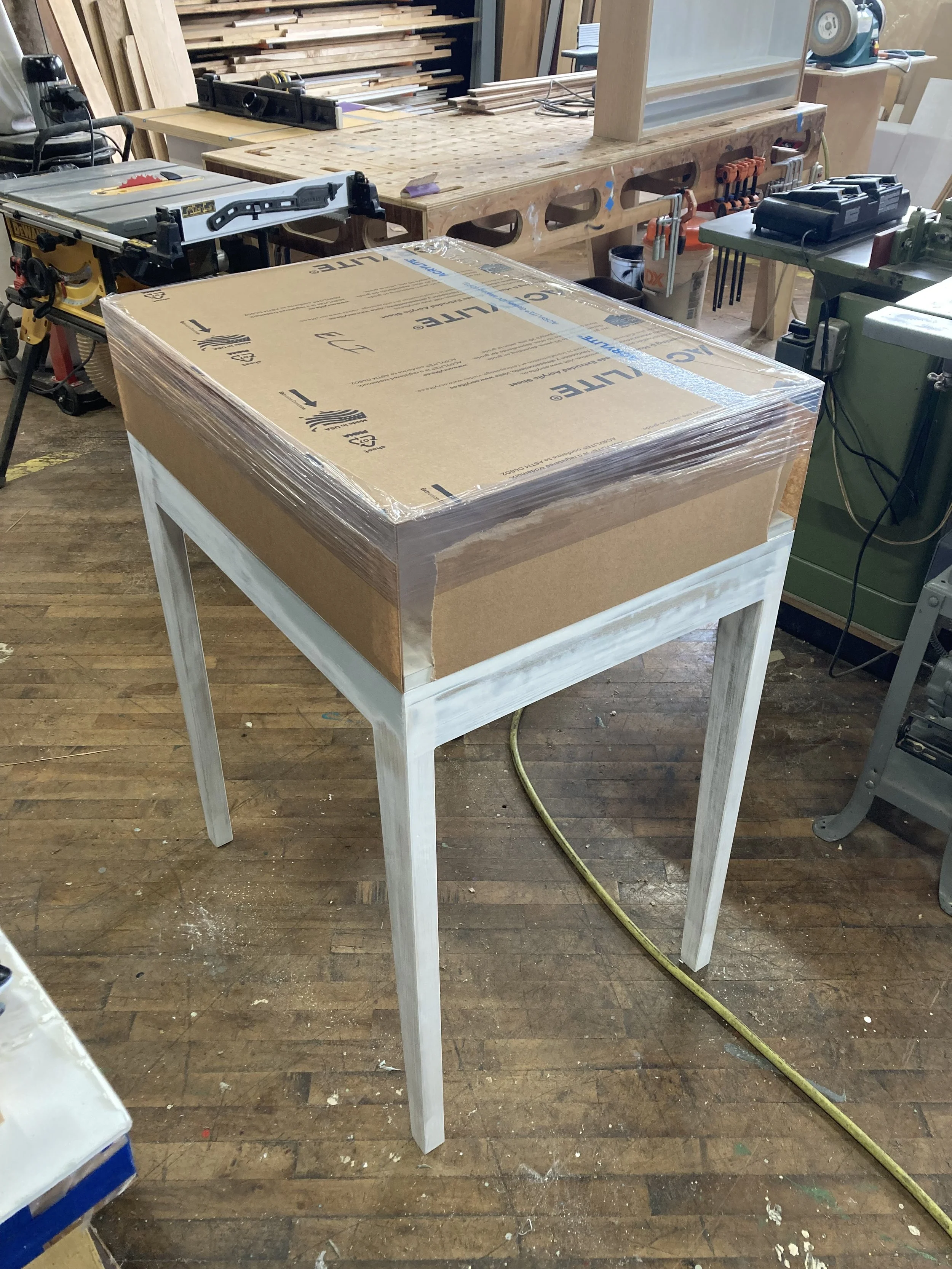Woodworking shop with a cardboard box wrapped in plastic atop a partially painted table, surrounded by various woodworking tools and equipment.