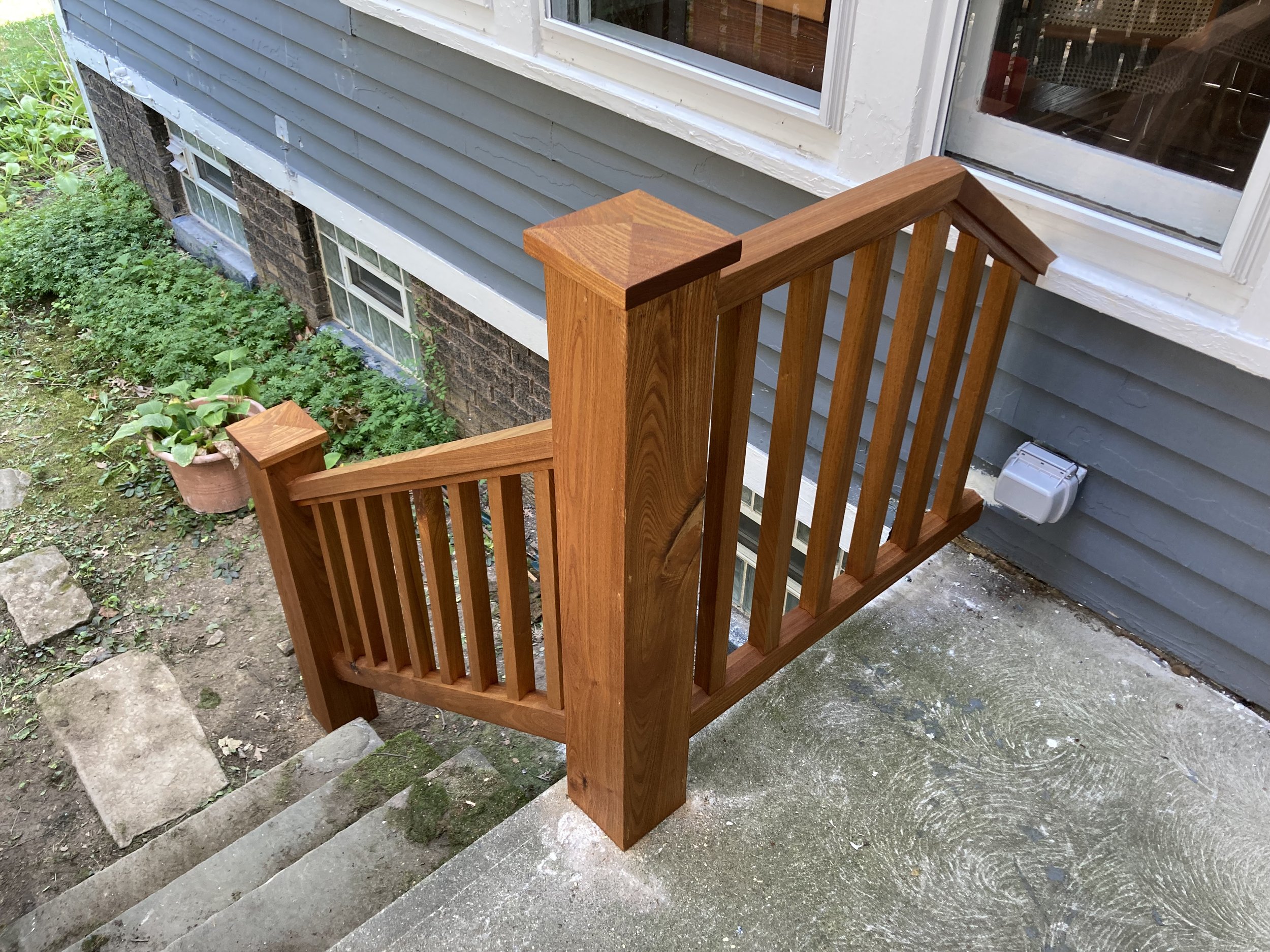 Wooden staircase railing leading to a gray house entrance with surrounding greenery.