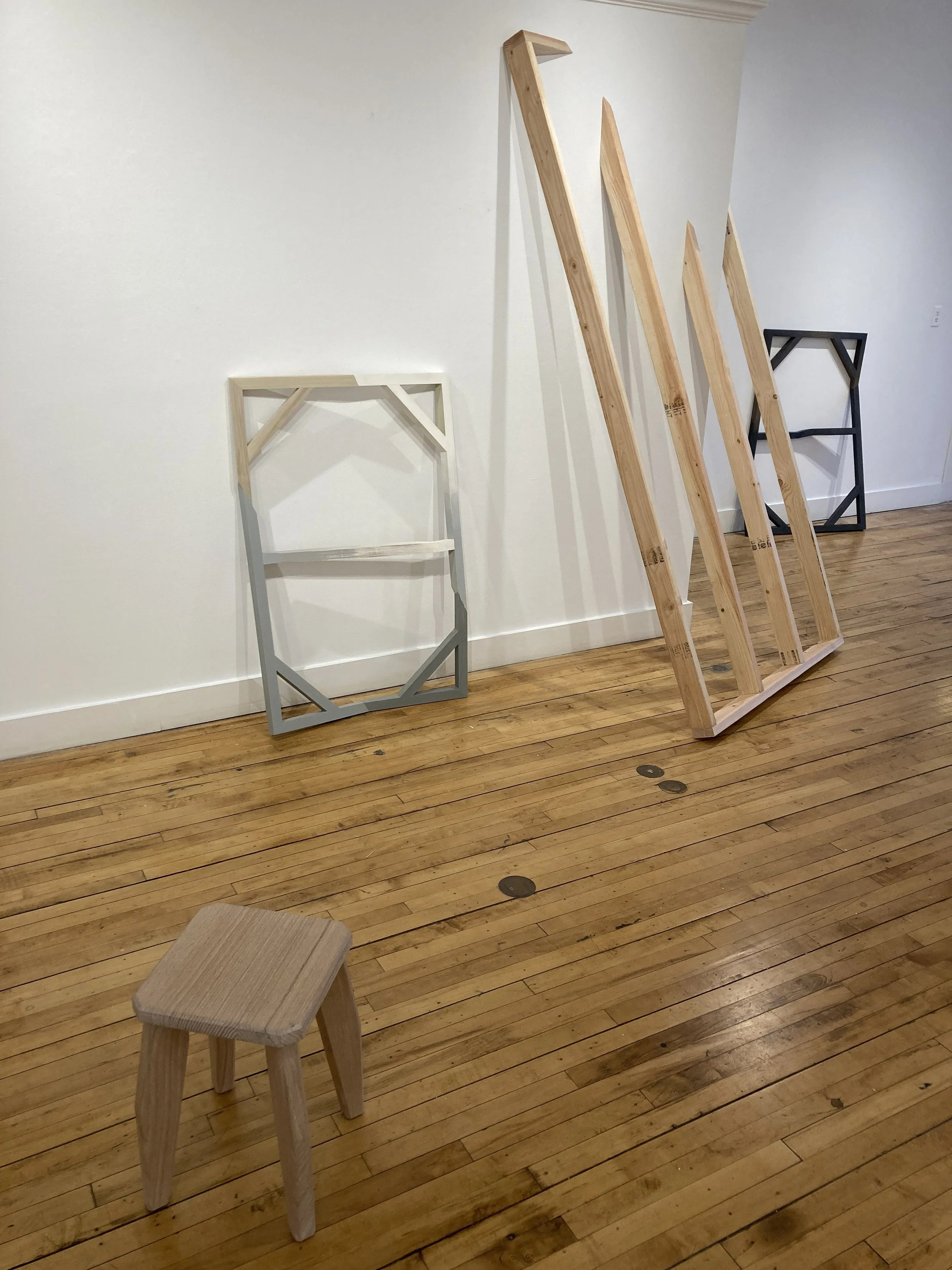 Wooden frames and structures leaning against a white wall, with a small wooden stool on a polished wooden floor.