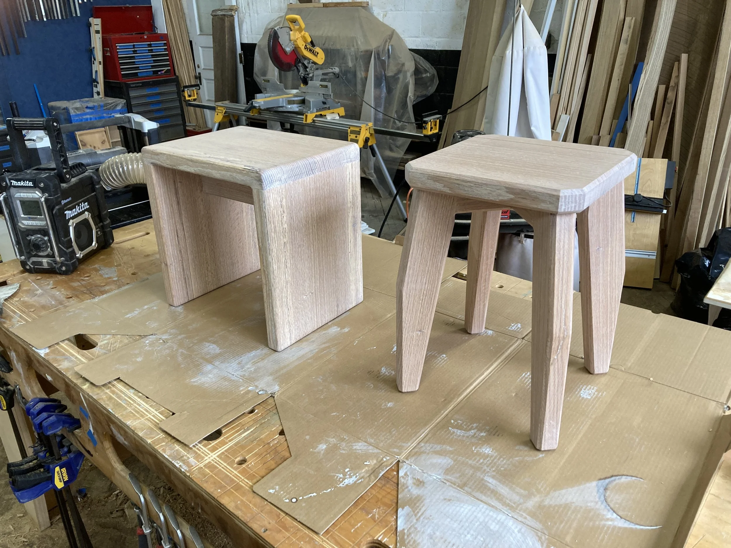 Two wooden stools on a workbench in a woodworking shop, surrounded by tools and materials.