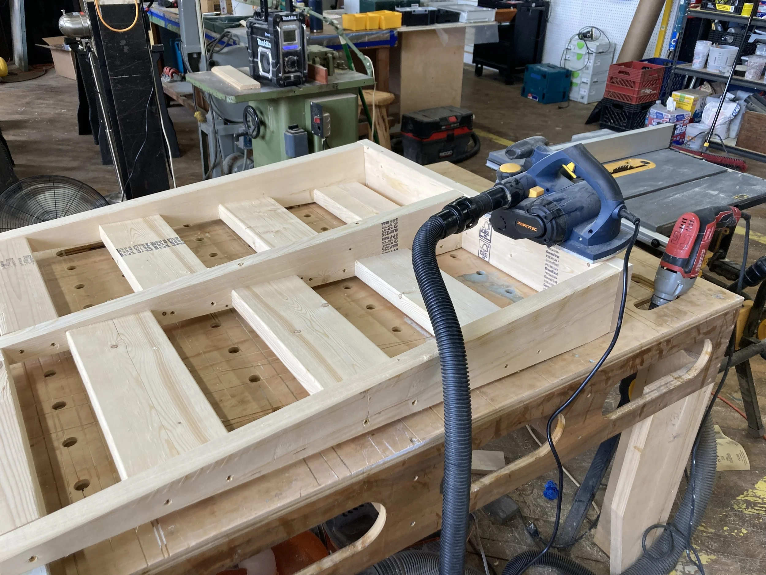 Workshop with wooden frame on workbench, power tools including a sander and drill, and various equipment in the background.