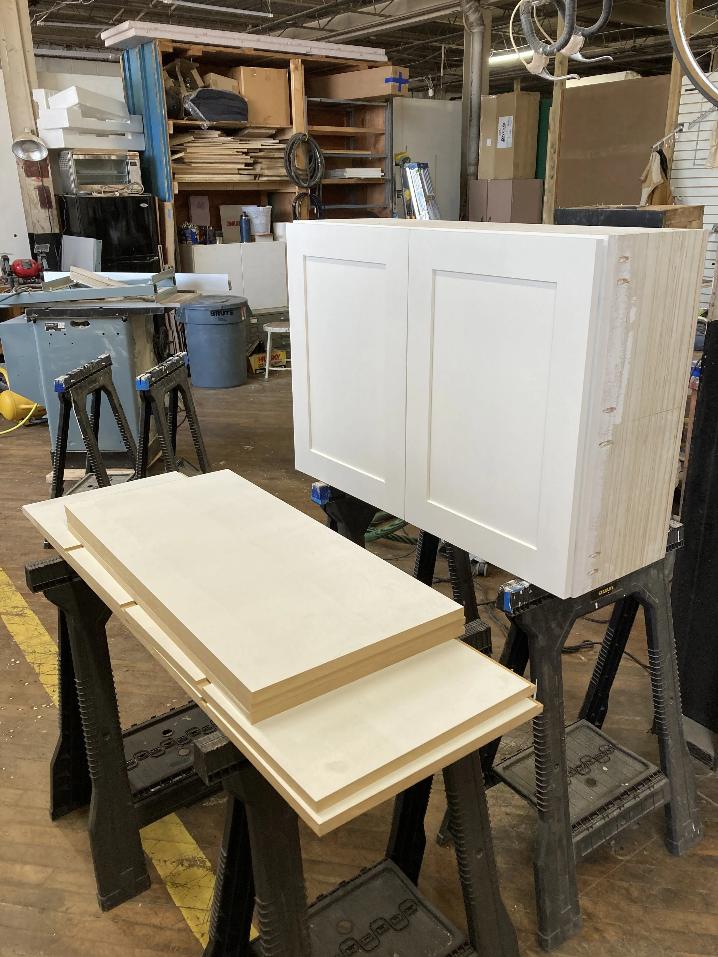 Woodworking shop with white cabinet doors, wooden planks on sawhorses, and various tools and materials in the background.