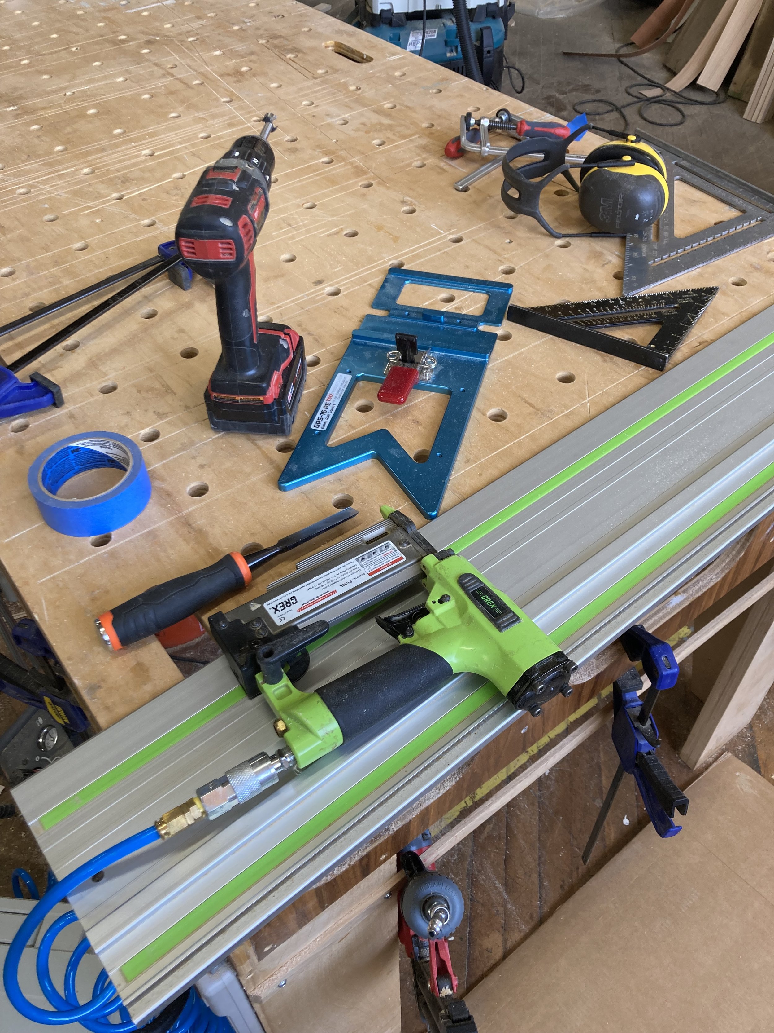 A workbench contains various tools and materials, including a cordless drill, measuring squares, a pneumatic nail gun, blue tape, and clamps. The surface is perforated and organized for woodworking purposes.
