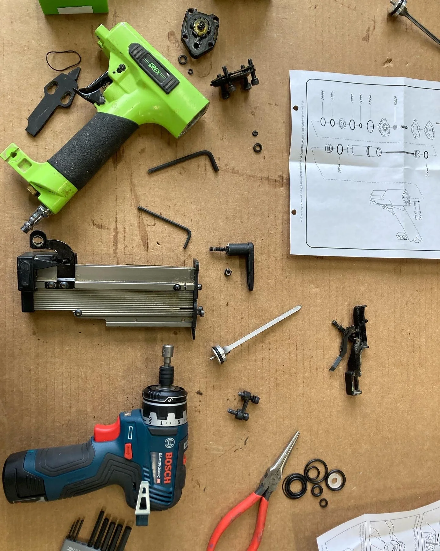 Disassembled nail gun with parts, drill, pliers, instruction manual on cardboard surface.