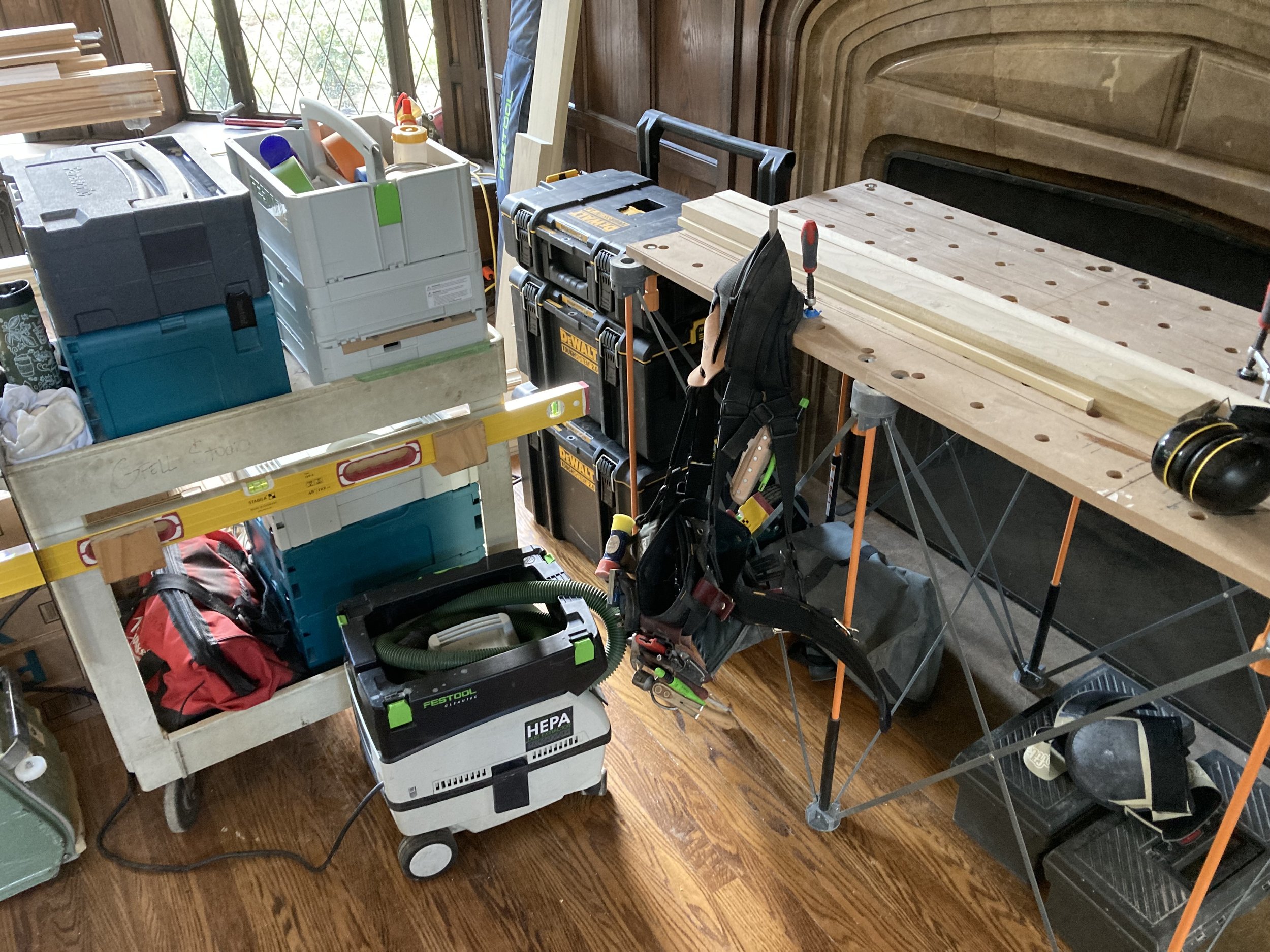 Woodworking workspace with tools and equipment. Includes a workbench, a tool cart, power tools, a vacuum cleaner, and wooden boards. Diverse tools and accessories are scattered around the area, with bright natural light coming in from the window.