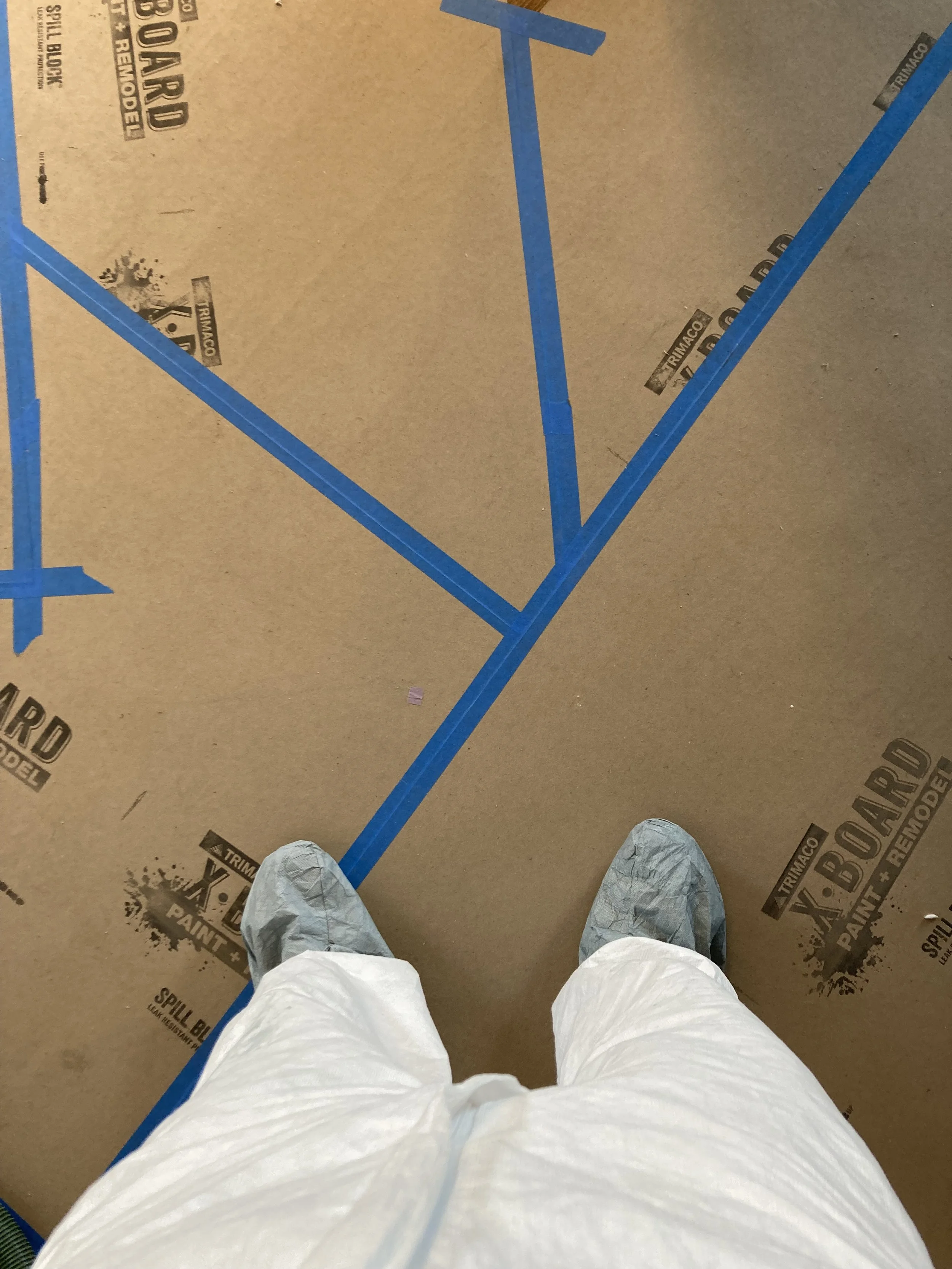 Person wearing shoe covers and a protective suit standing on cardboard covered floor with blue tape markings.