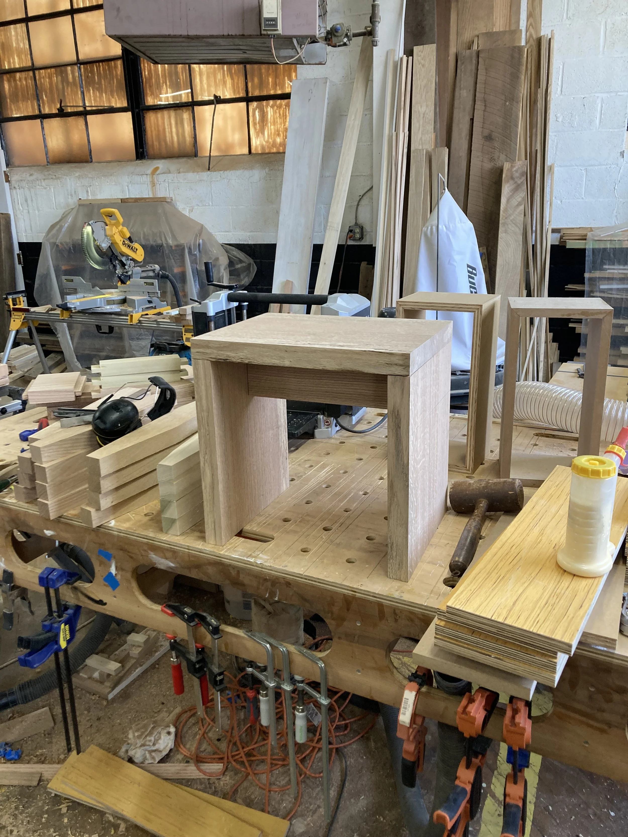 Woodworking workshop with a workbench, a miter saw, wooden planks, clamps, and an unfinished wooden stool.