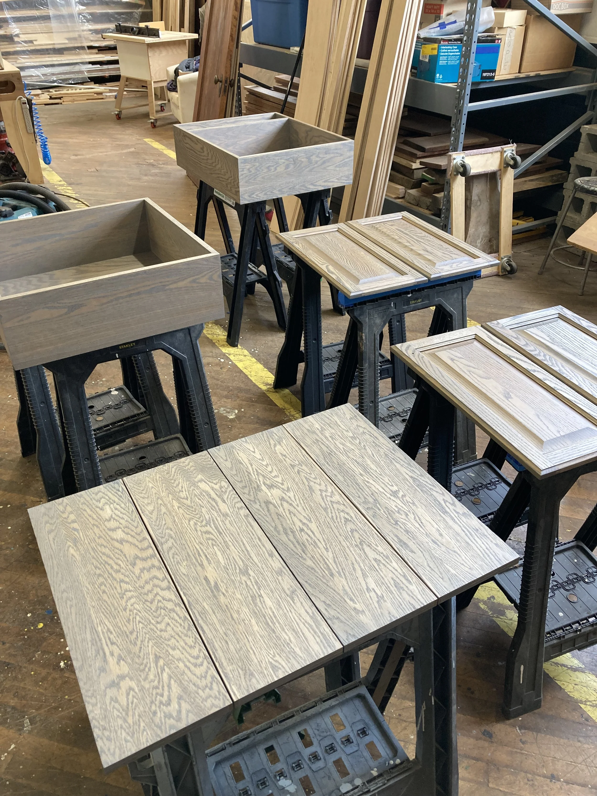 Woodworking shop scene with saw horses supporting wooden boxes and panels, including drawer fronts and cabinet components.