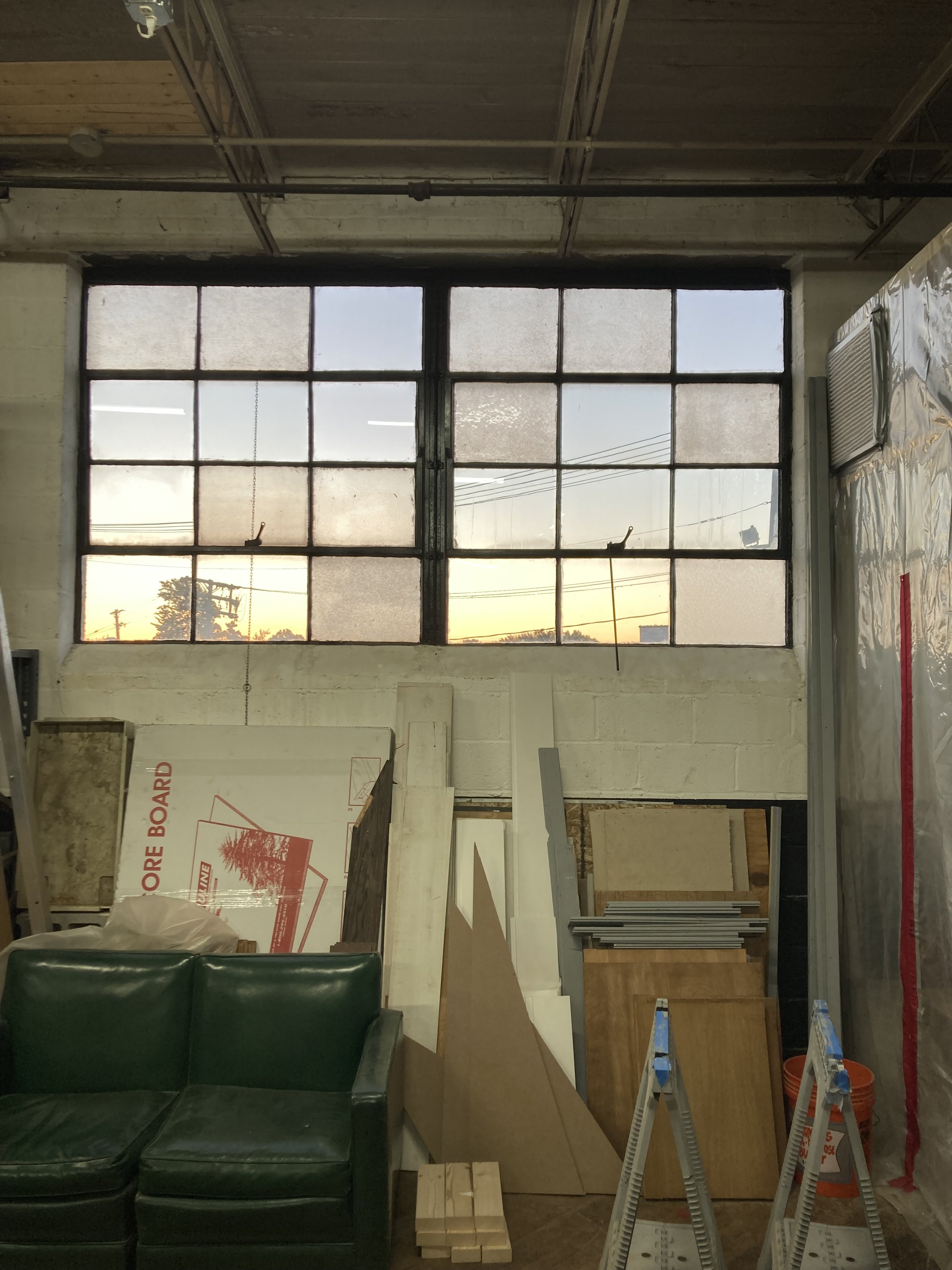 Interior of a workshop with large industrial windows, wooden boards stacked against a wall, a green leather couch, and scaffolding frames with buckets.