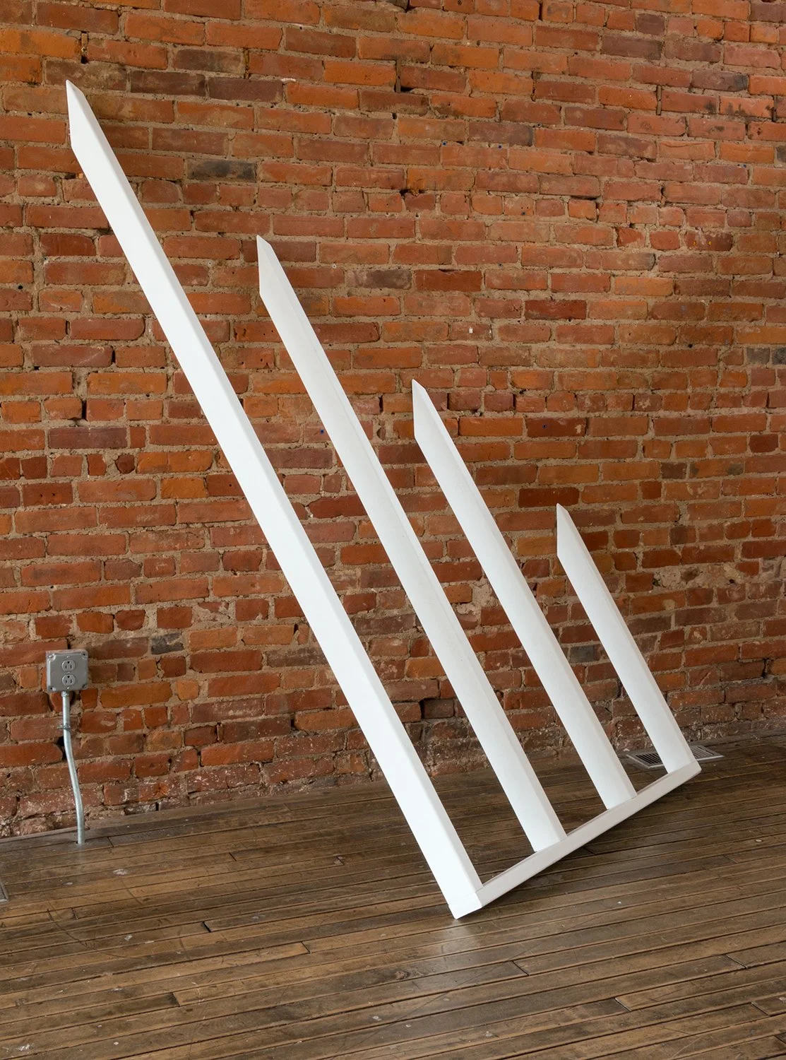 White angular sculpture against exposed brick wall on wooden floor.