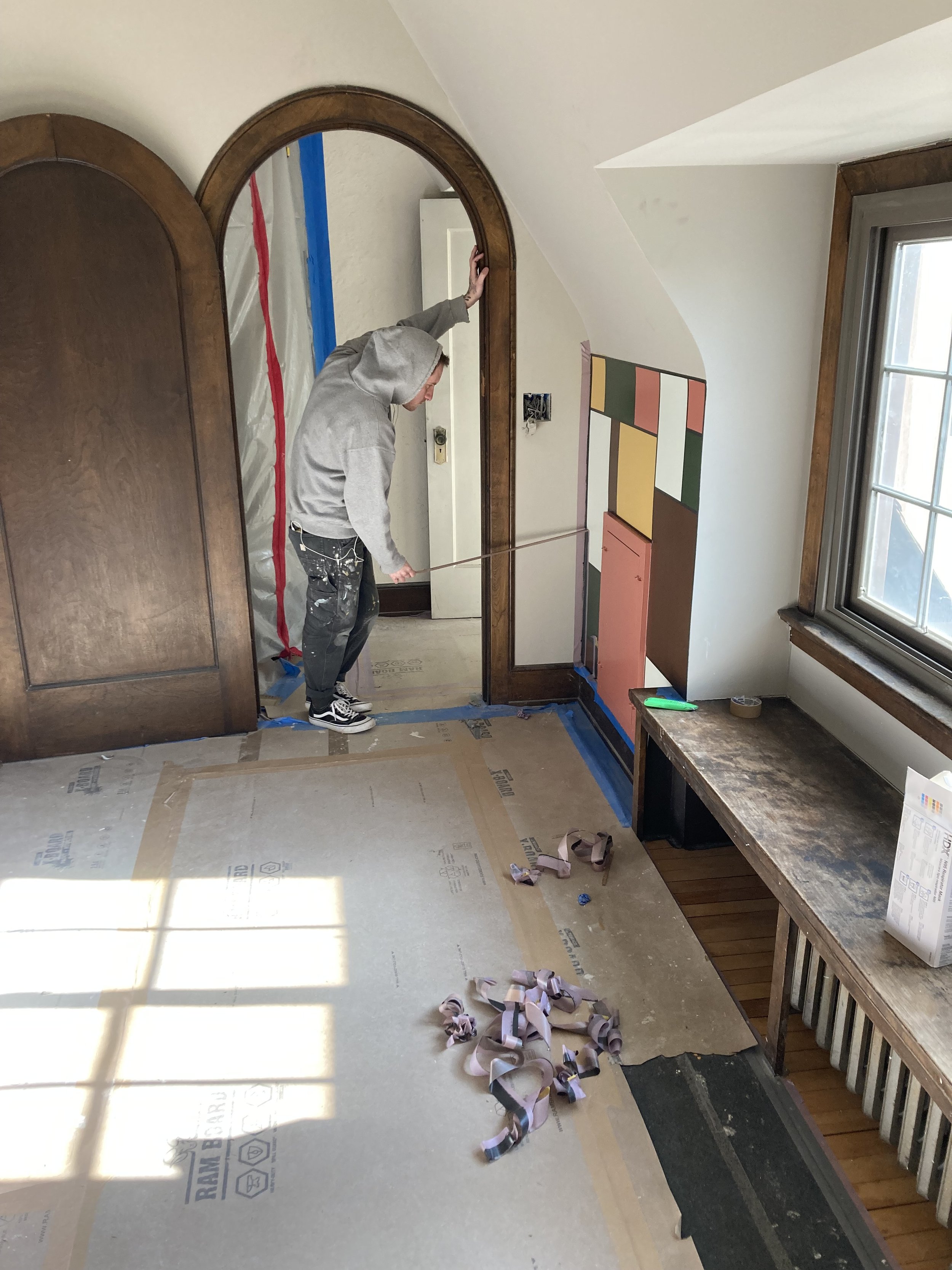 Person in a gray hoodie working on a room renovation around a large wooden arch, with protective materials on the floor and walls, and colorful paint samples visible on the wall.