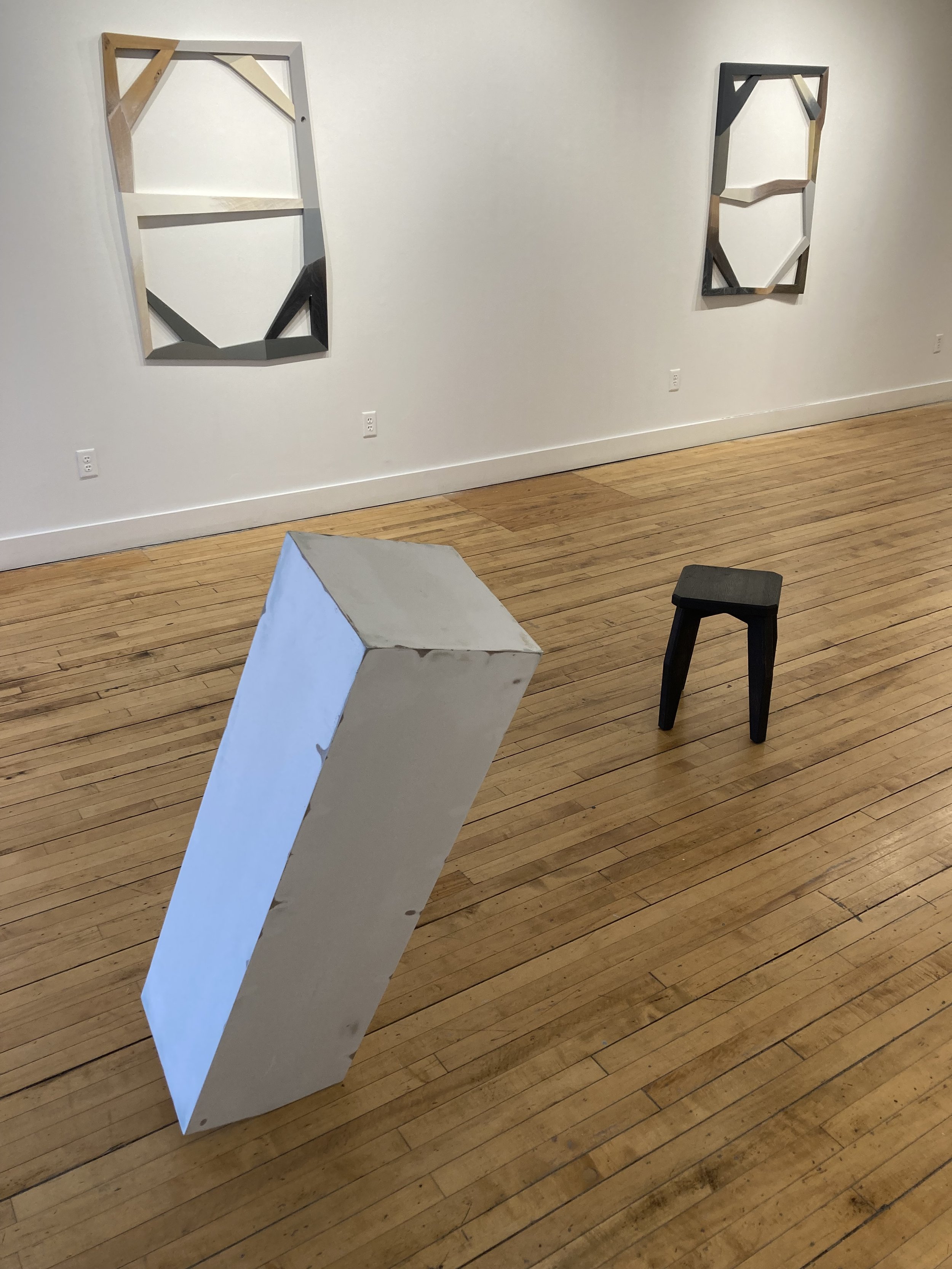 Contemporary art gallery with abstract geometric wall sculptures, a leaning rectangular object, and a small black stool on a wooden floor.