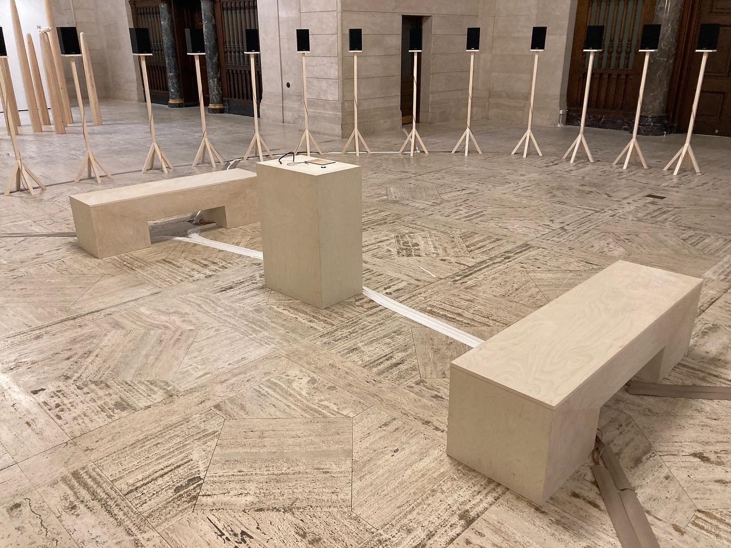 Art installation featuring wooden benches and multiple speakers on stands in a spacious room with marble flooring.