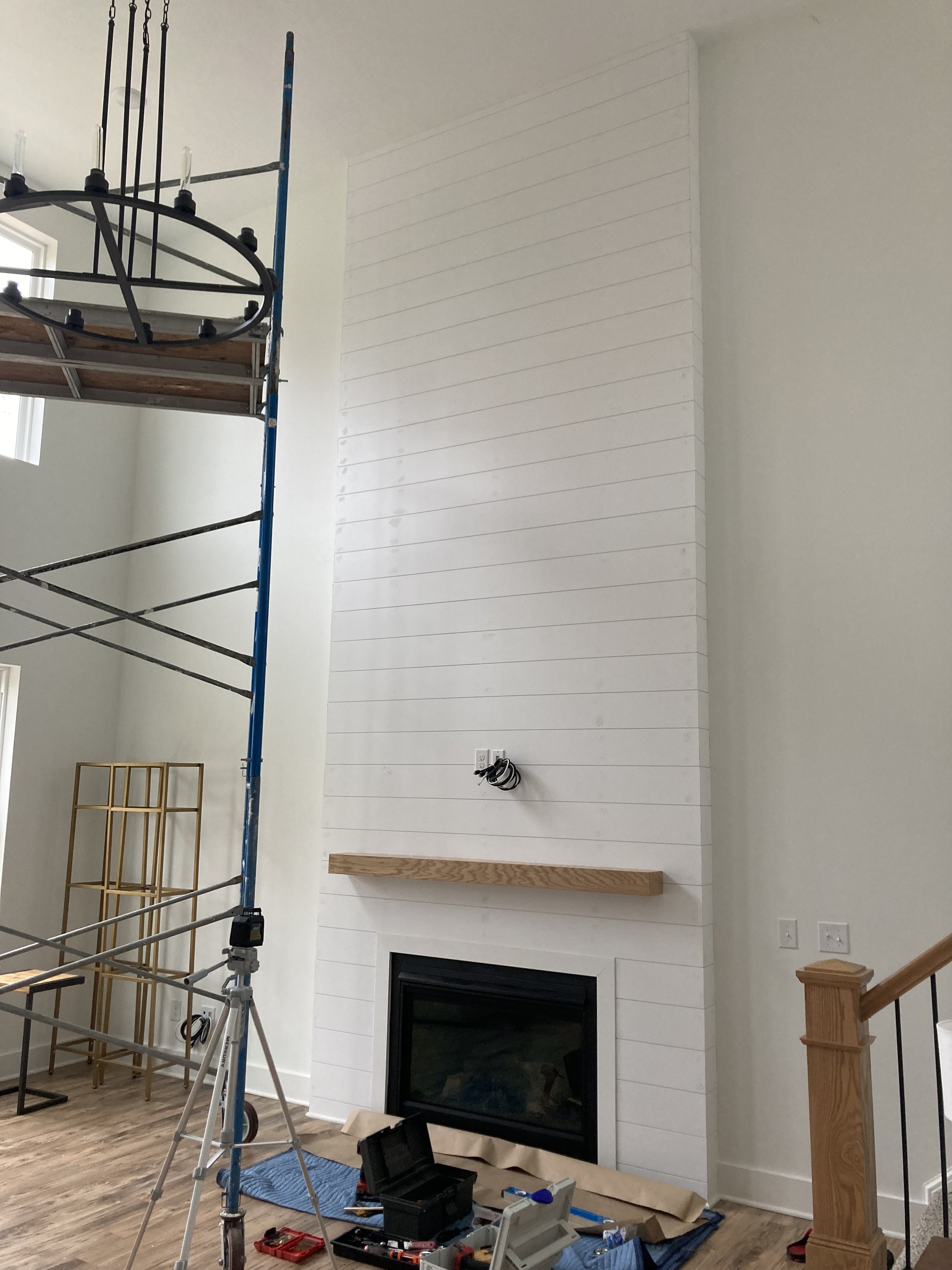 Interior room with a tall white shiplap fireplace, wooden mantel, and construction tools. Scaffolding supports a light fixture. A staircase with wooden railing is visible on the right.