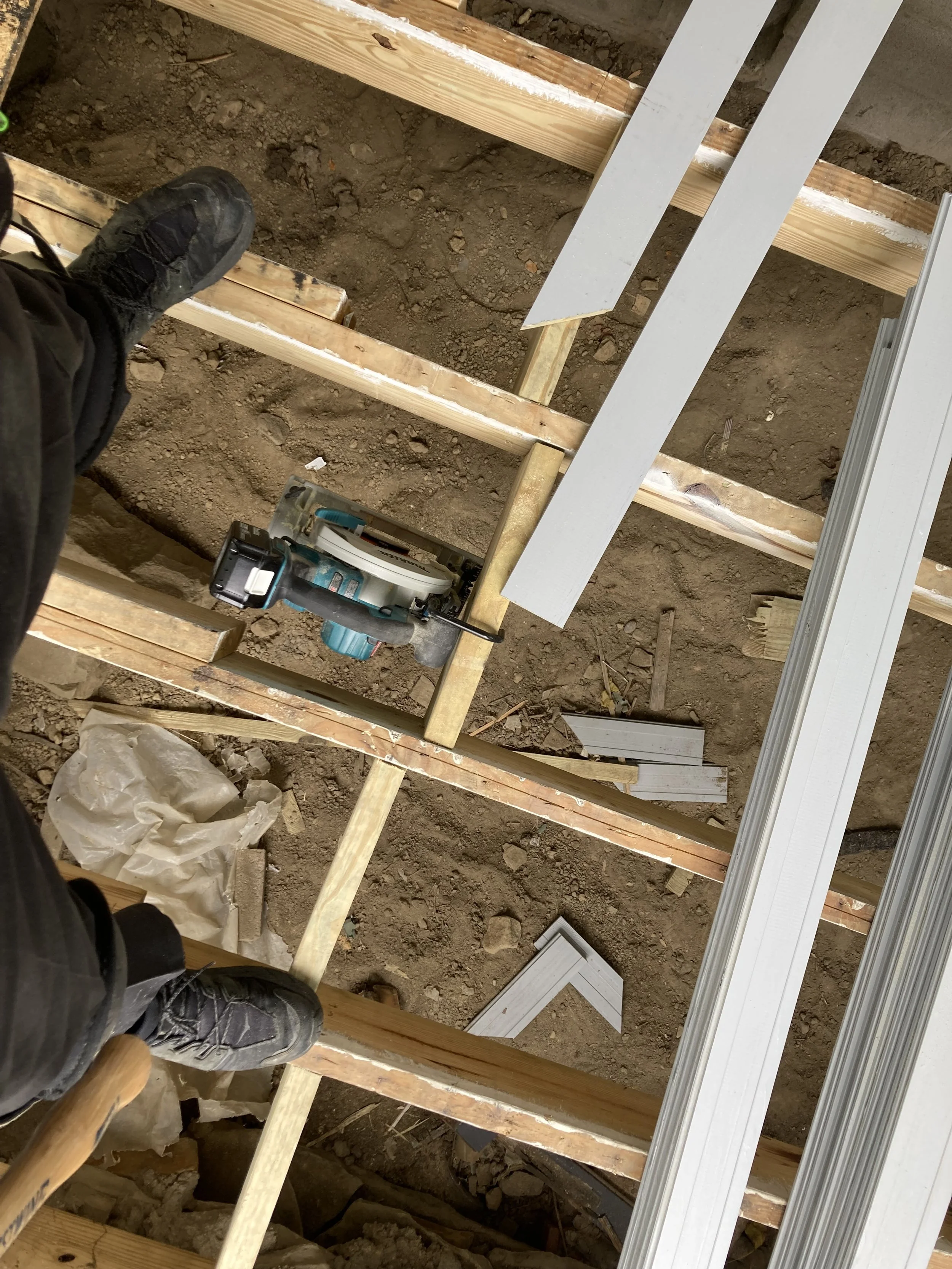 A construction site showing wooden floor joists, a circular saw, long white boards, and a person wearing black shoes standing on the beams.