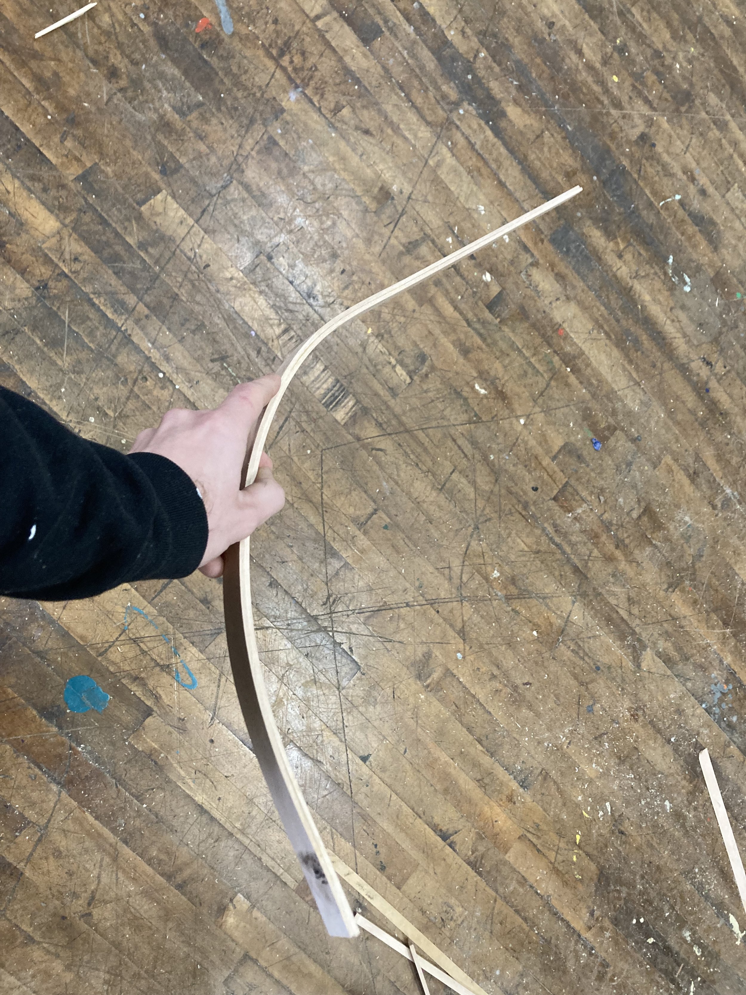 Person holding a curved piece of wood, possibly a thin plywood strip, against a wooden floor with scattered paint stains.