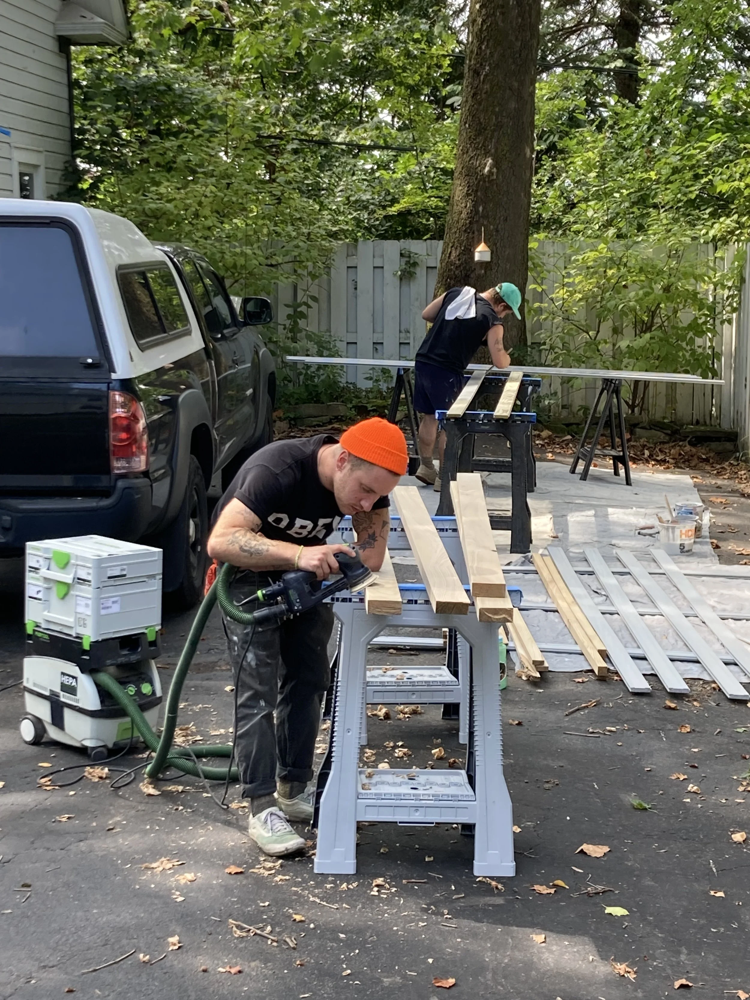Two people woodworking in a driveway, sanding wood planks on sawhorses with a sander, near a vehicle and surrounded by trees.