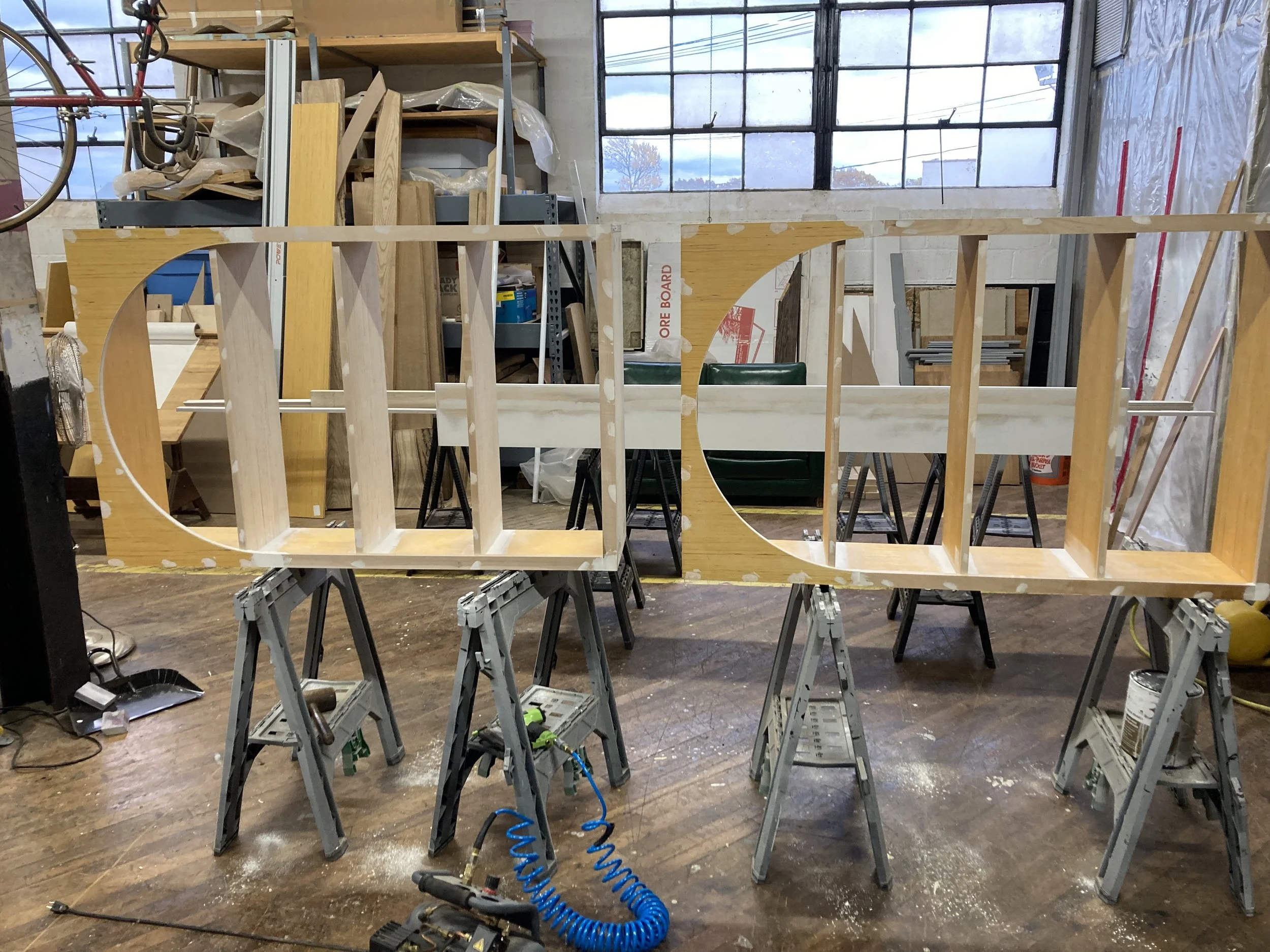 Wooden structure being constructed, supported by sawhorses in a workshop with various tools and materials in the background.