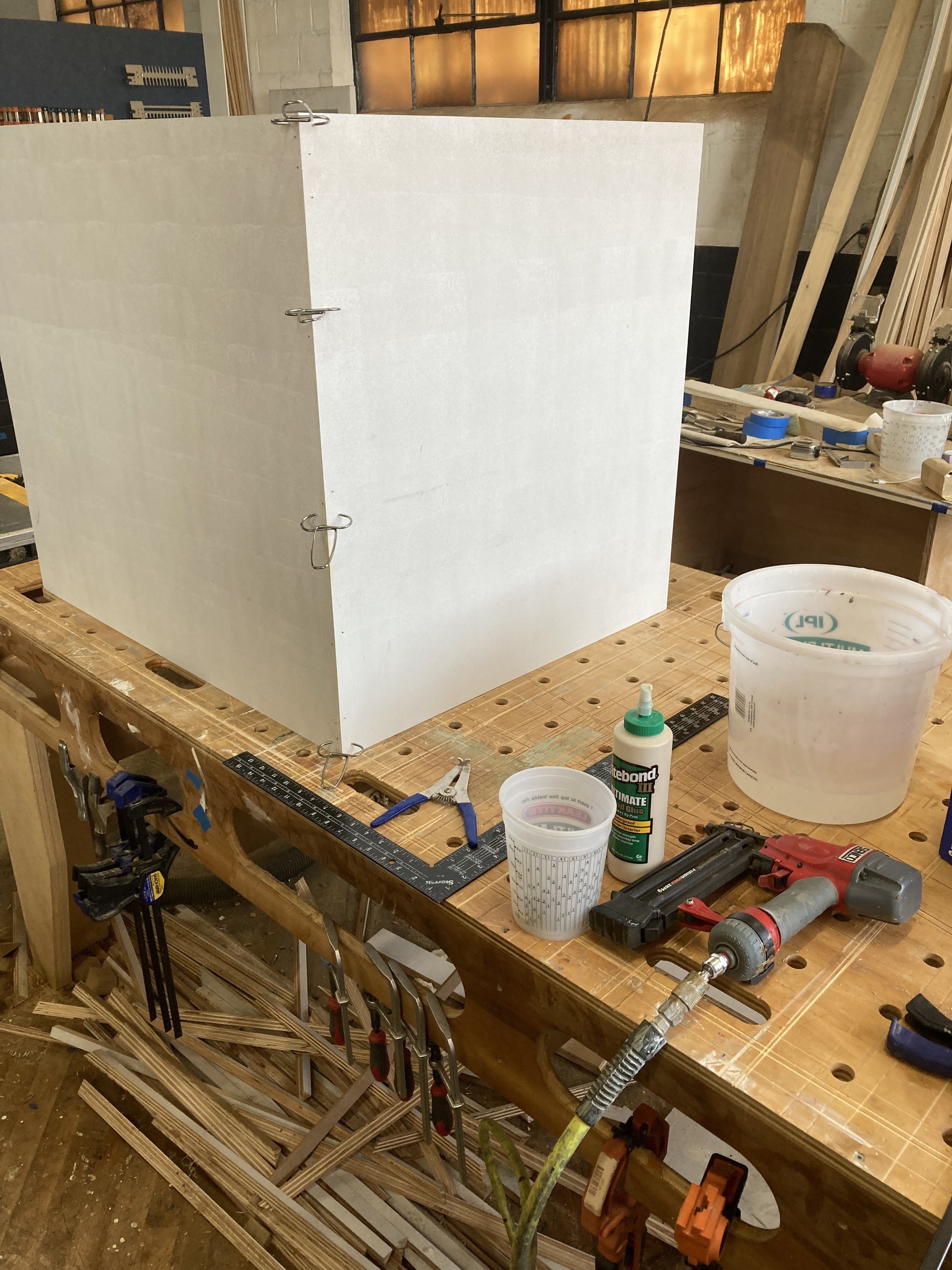 Woodworking bench with large white box, clamps, glue, tools, and wood scraps in a workshop.