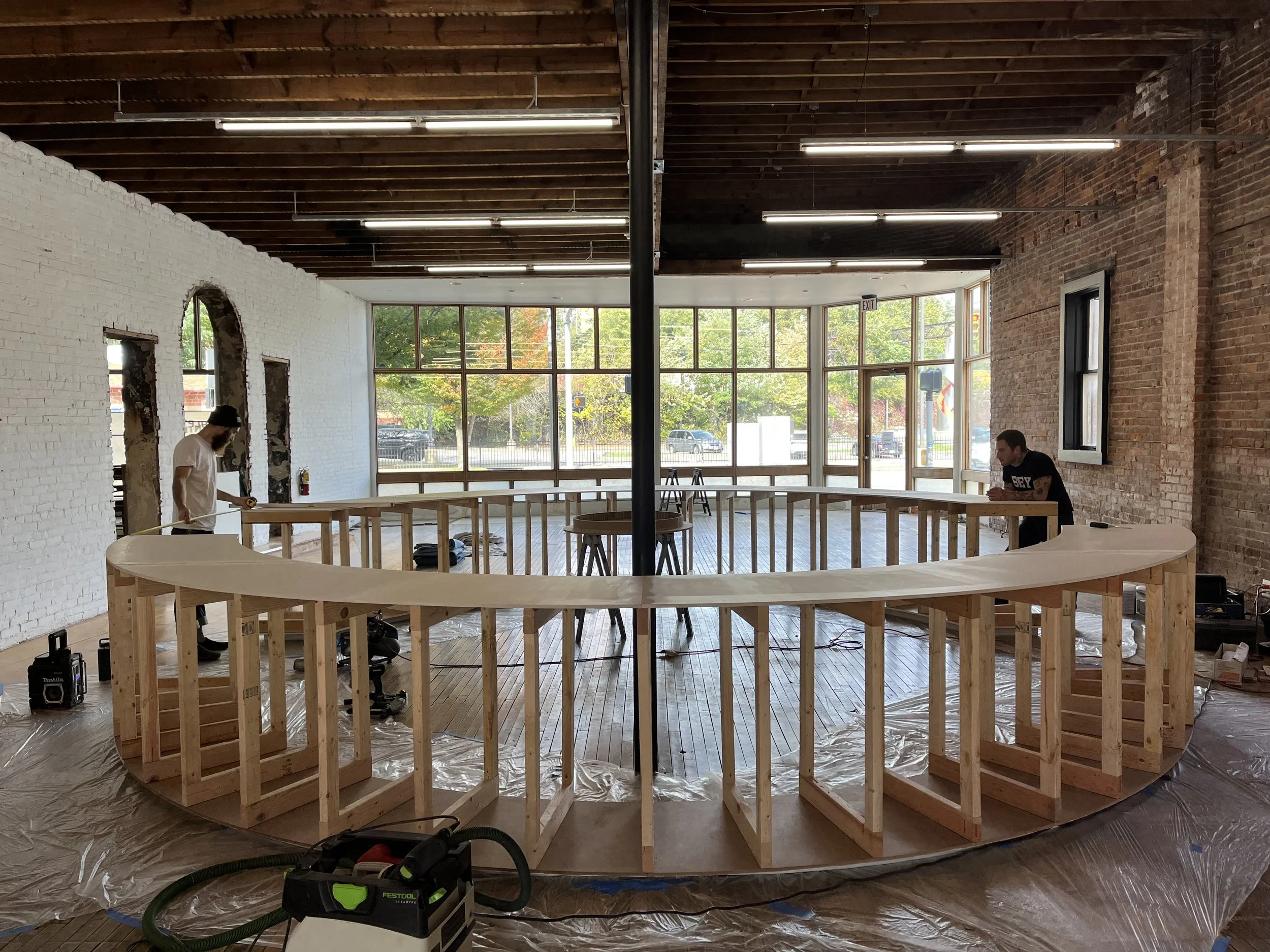 Two people constructing a large circular wooden frame inside a room with exposed brick walls and large windows, under a wooden ceiling with fluorescent lights.