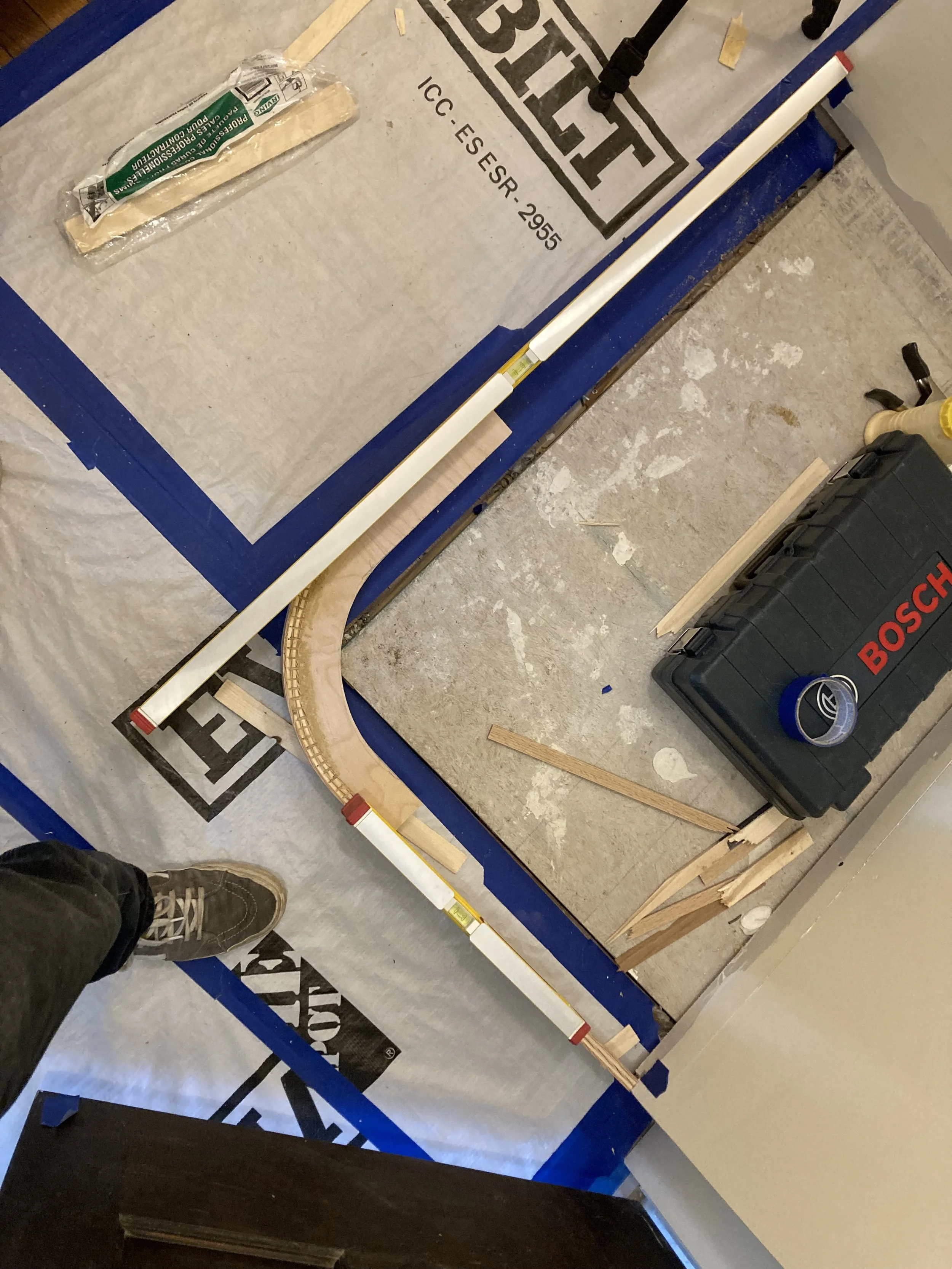 Construction tools and materials on paper-covered floor, including a Bosch toolbox, wooden planks, tape, and measuring tape.