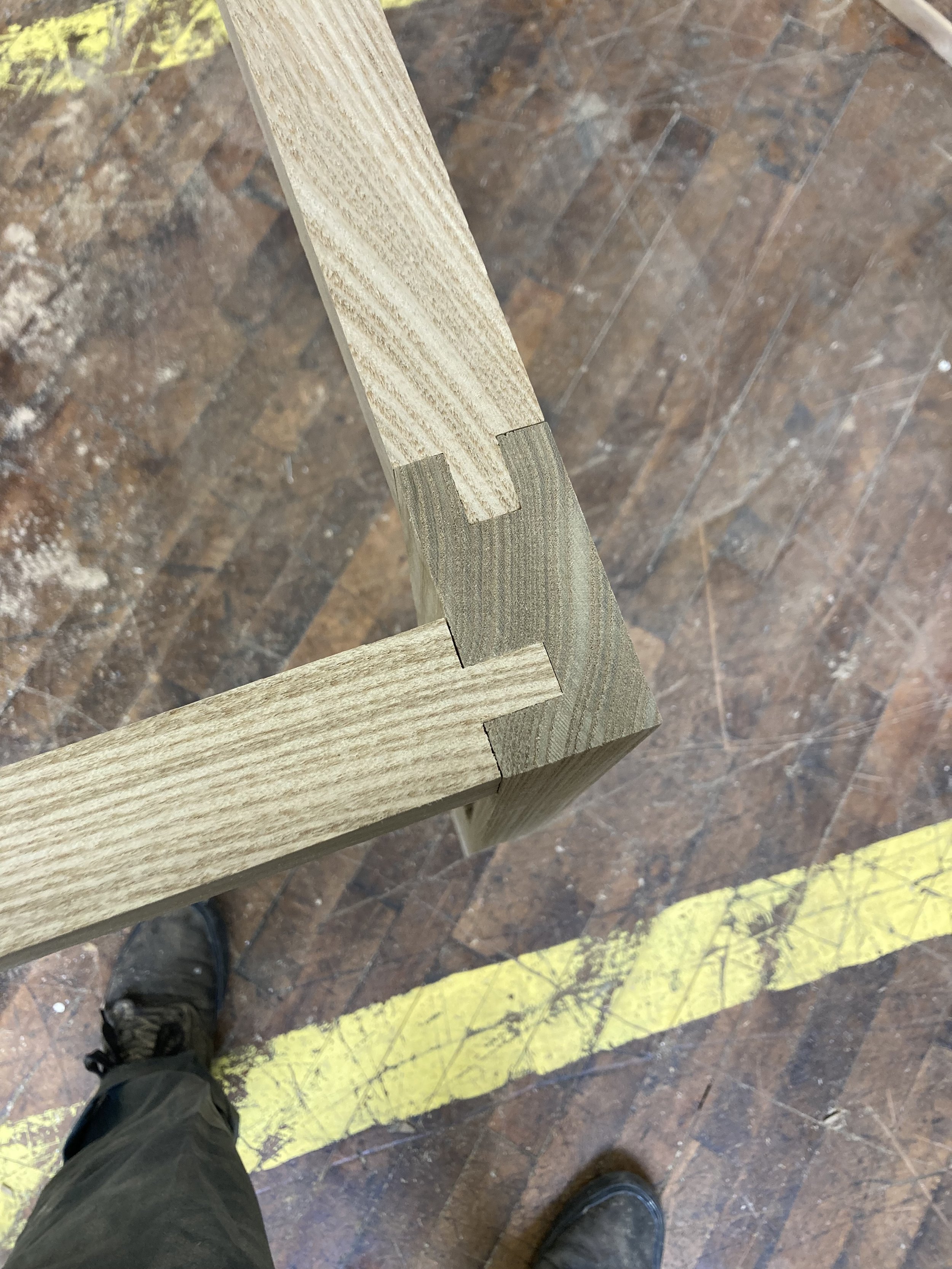 Corner view of a wooden frame with a finger joint, over a worn wooden floor with yellow tape, showing a person's foot.
