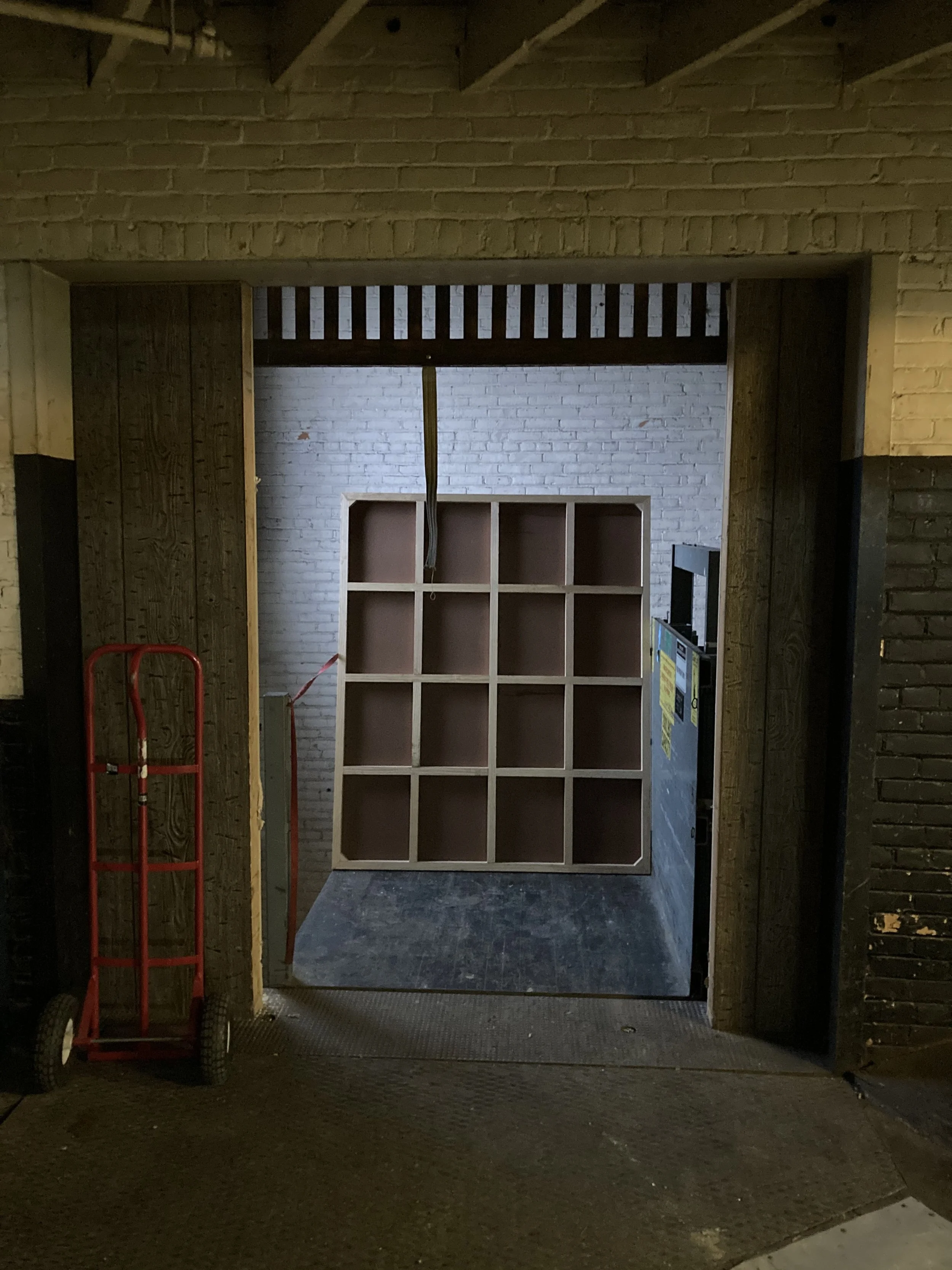 Elevator opening with a dolly cart and a large shelving unit inside, white brick walls visible.