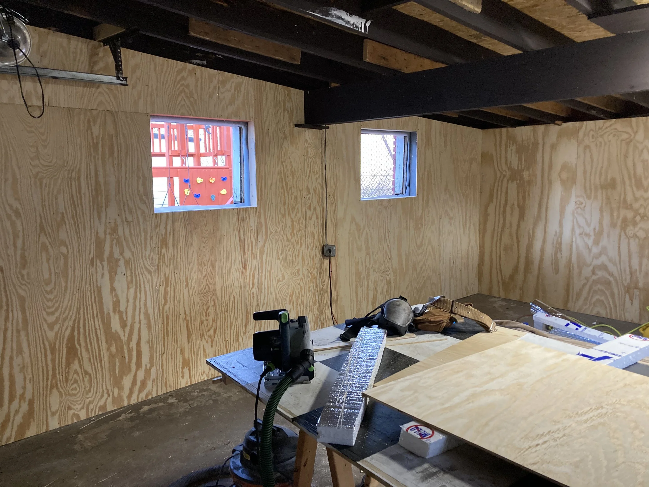 Interior of a room under construction with plywood walls, a workbench with tools, and two small square windows.