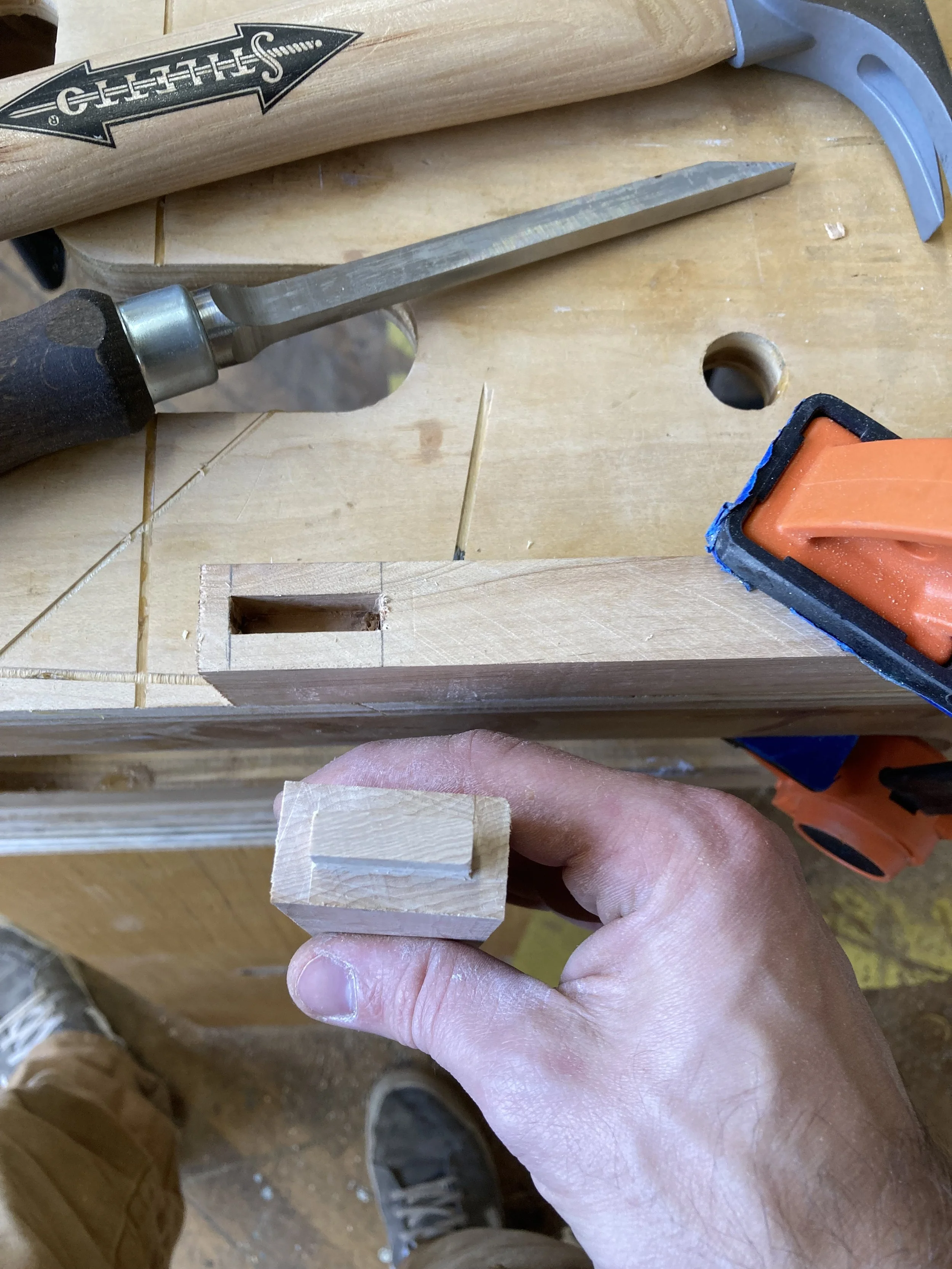 A person holding a wooden tenon in front of a partially completed mortise on a workbench, with a chisel and hammer nearby.