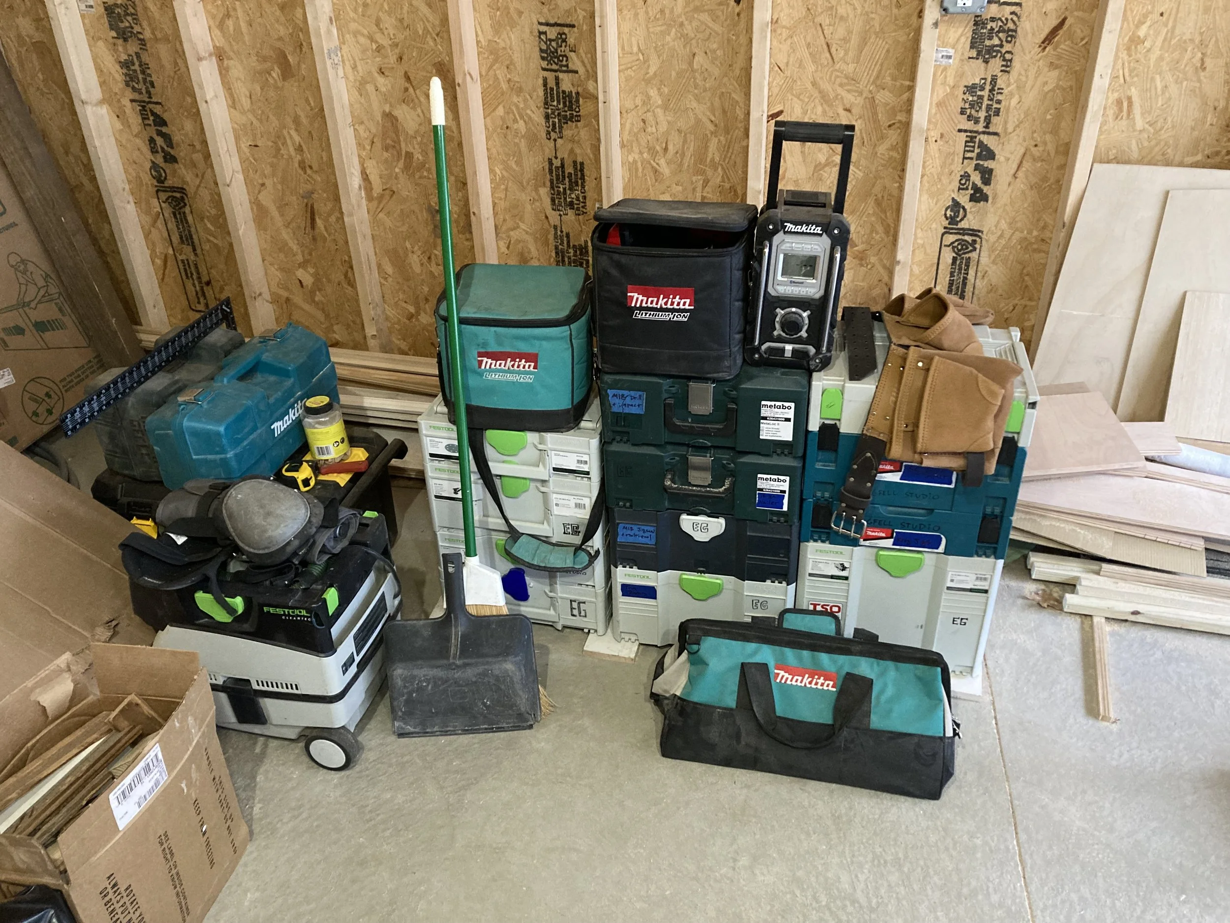 Construction tools and equipment stacked against a wooden wall, including Makita tool bags, a Festool vacuum, power tool cases, a broom, a shovel, and plywood sheets.