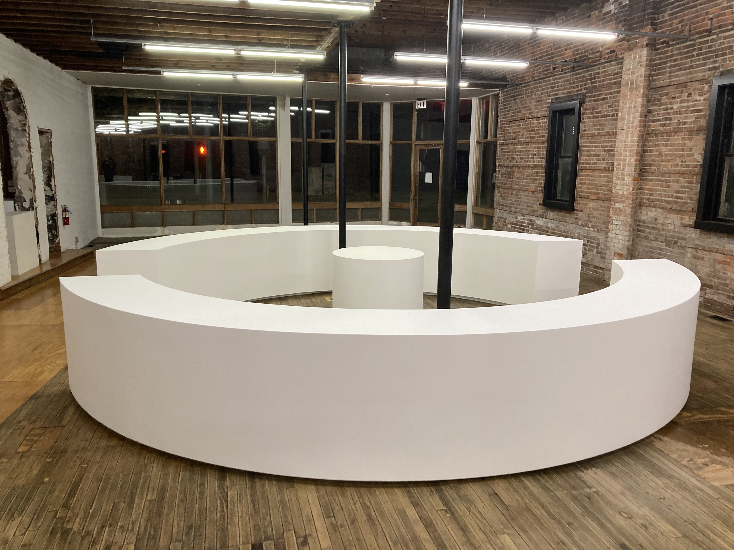 Modern office lobby with curved white desks arranged in a circular formation, exposed brick walls, wooden ceiling with fluorescent lights, and large windows reflecting ceiling lights. The floor is wooden, and there is a central black pole.