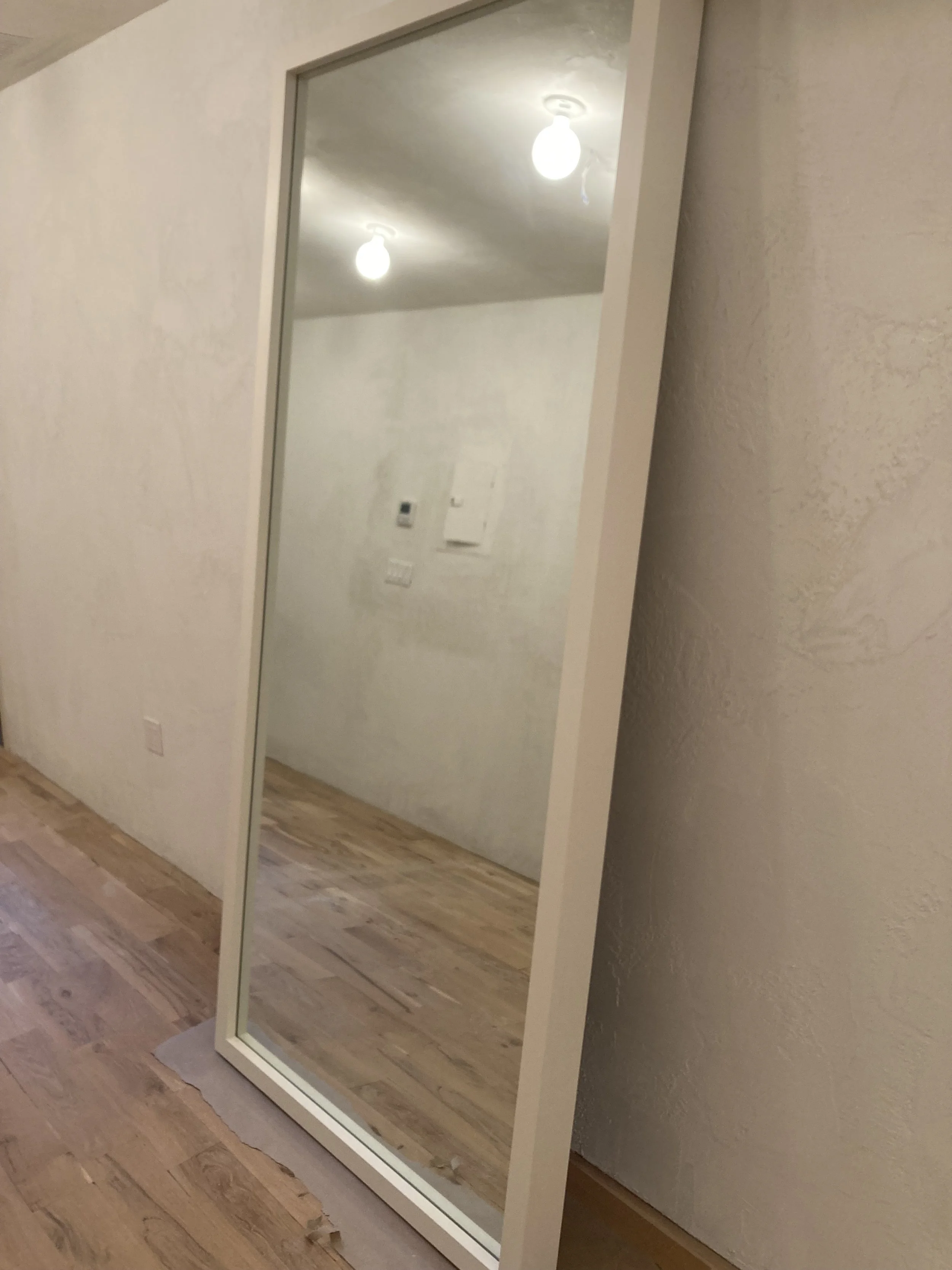 Large framed mirror leaning against a textured wall in a room with wooden flooring and bright ceiling lights.