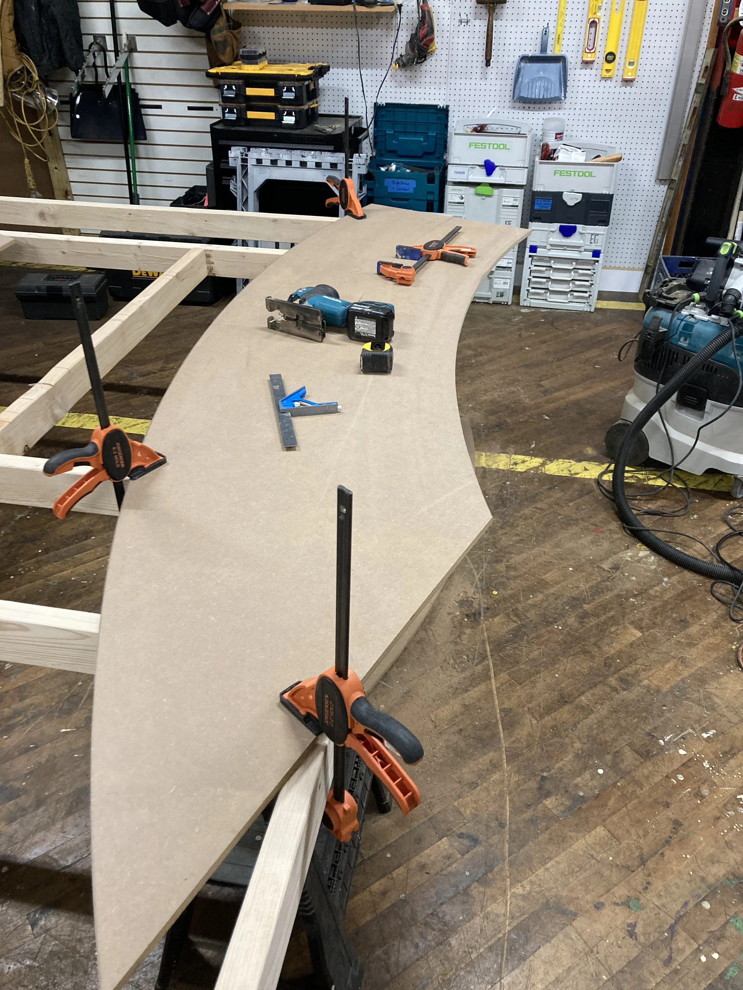 Woodworking workshop with a curved wooden piece clamped to a workbench, various tools and equipment visible, such as a jigsaw, clamps, and boxes.