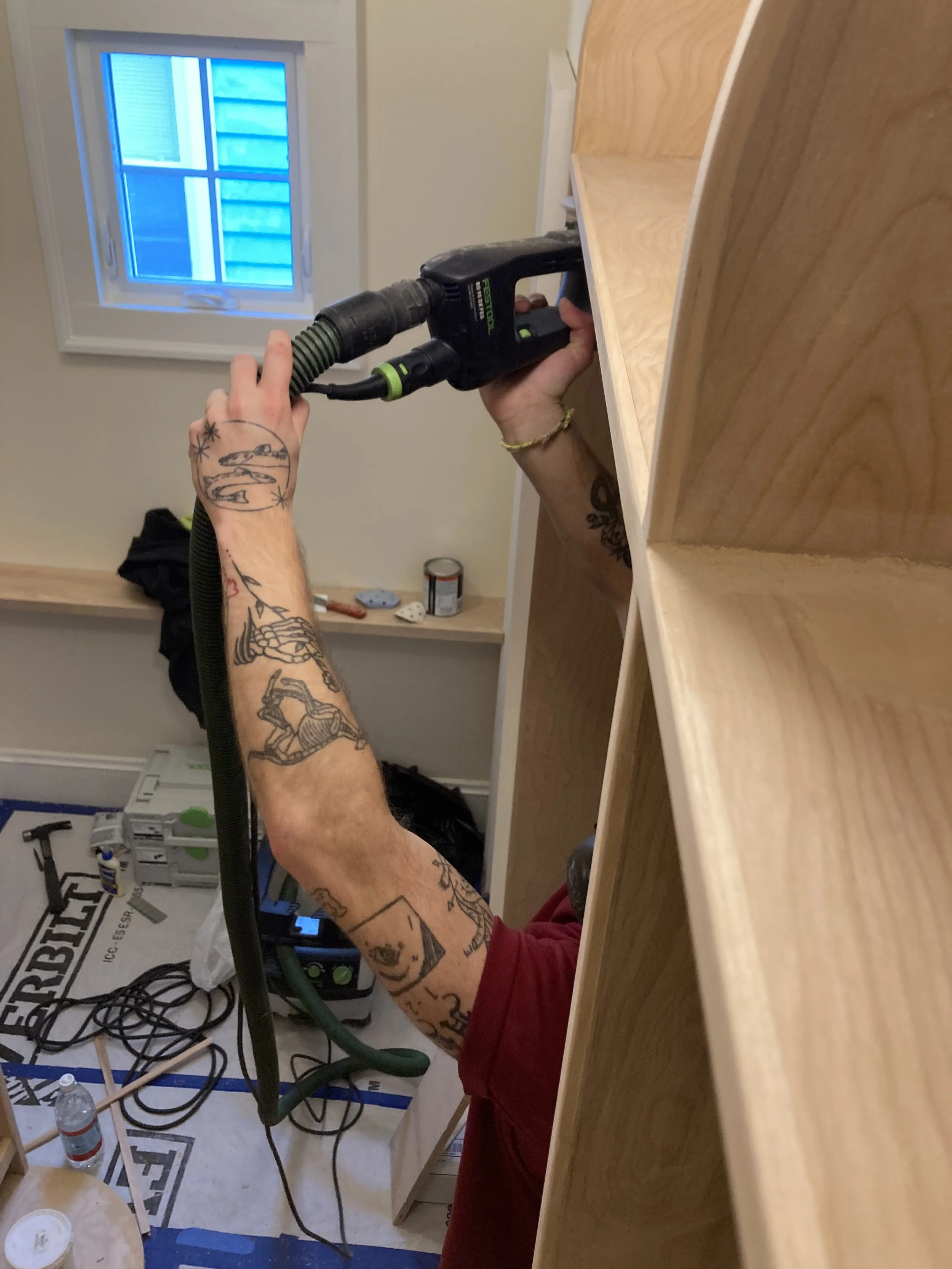 Person using a power drill on a wooden structure in a workshop. Visible arm has tattoos. Tools and materials are scattered on the floor.