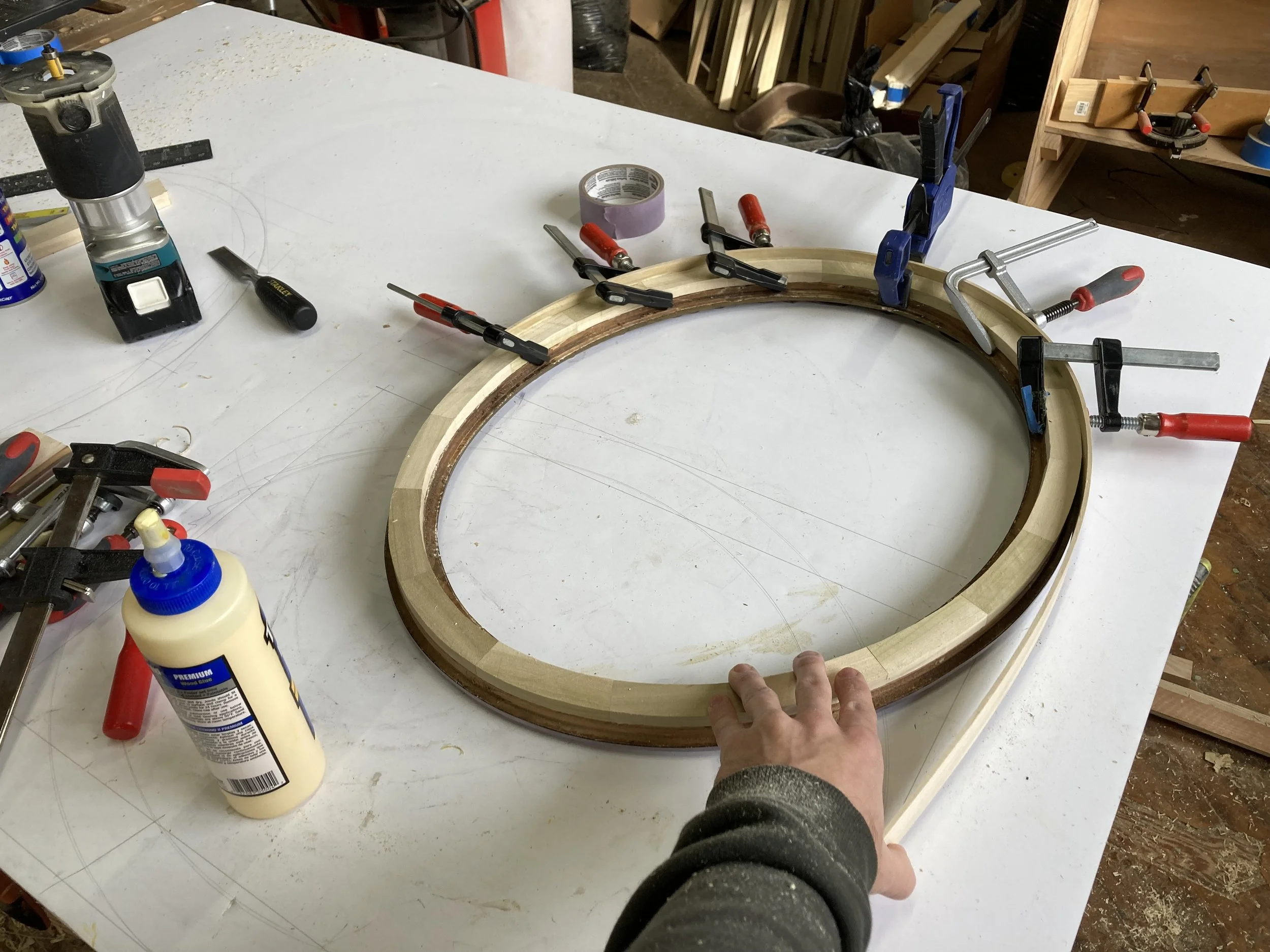 Woodworking clamp setup for a circular frame on a workbench, surrounded by tools like a glue bottle, router, clamps, and chisel in a workshop.