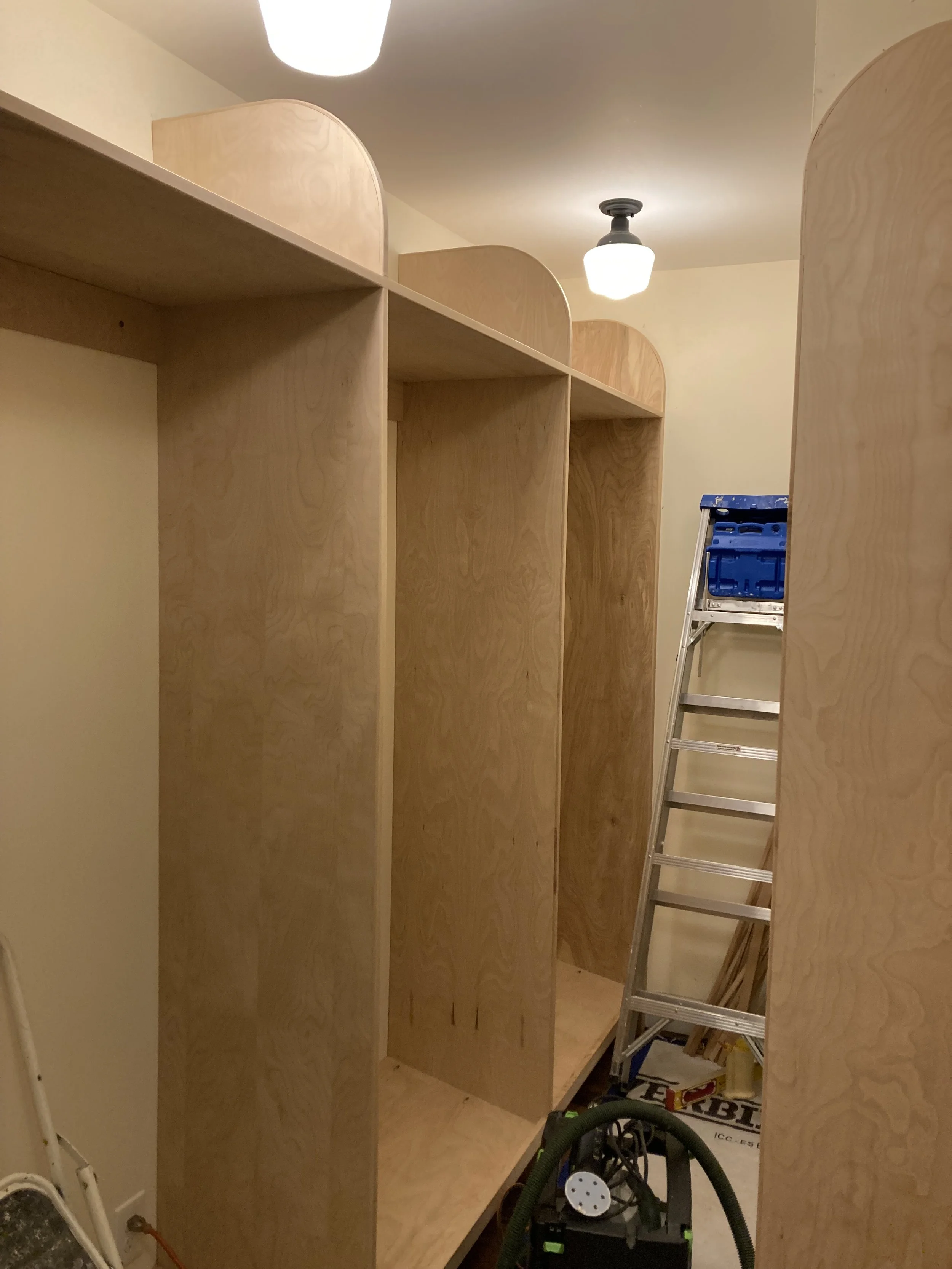 Wooden closet compartments under construction, with a ladder and tools in a room with overhead lighting.