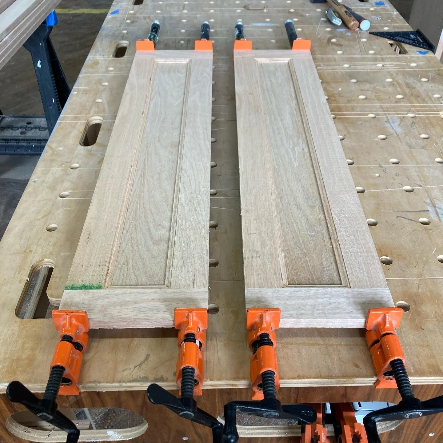 Two wooden panels clamped on a workbench with orange clamps, in a woodworking shop.